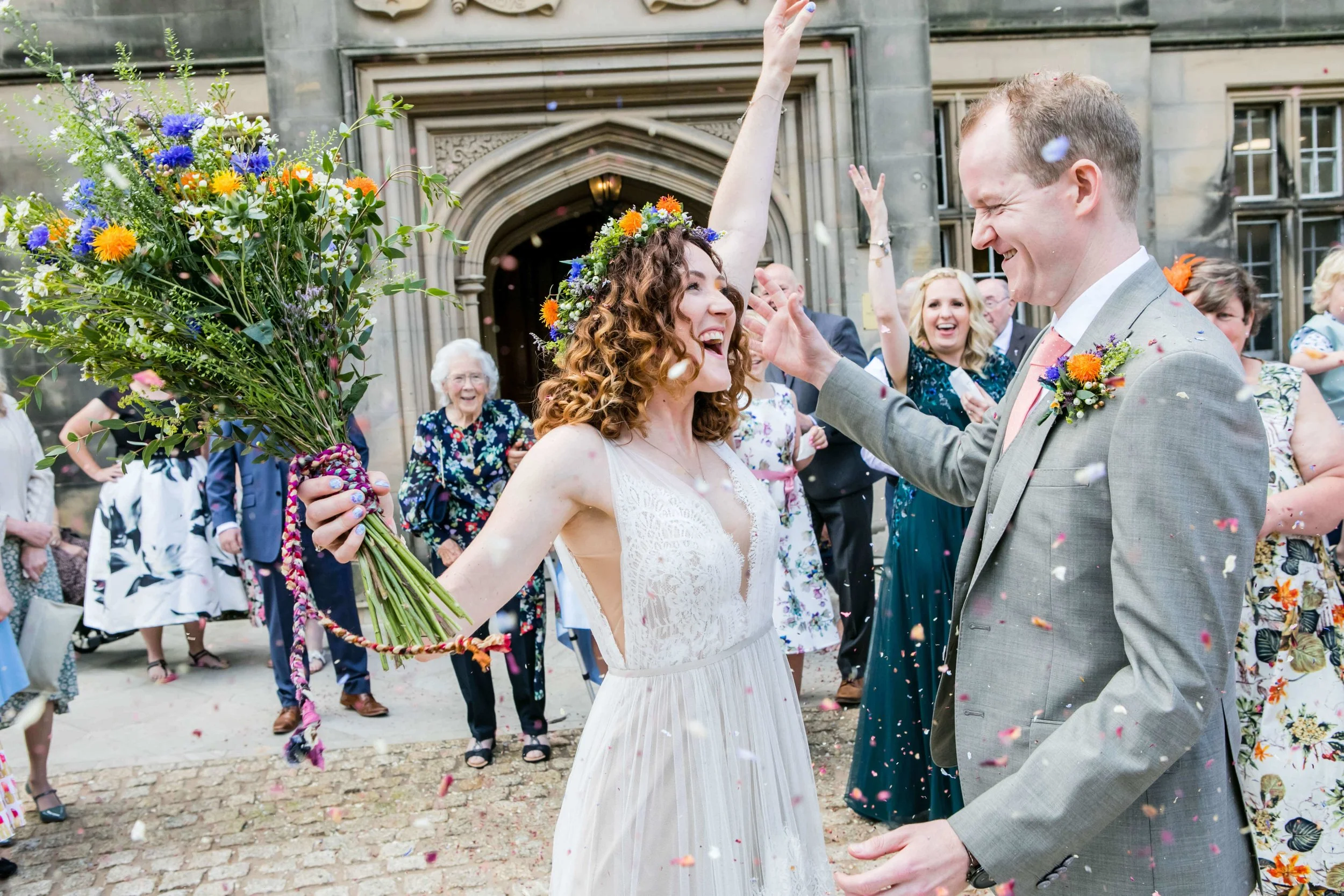 A bride and groom celebrating their wedding outside Matfen Hall with friends and family, confetti falling, the bride holding a large bouquet of colourful flowers.