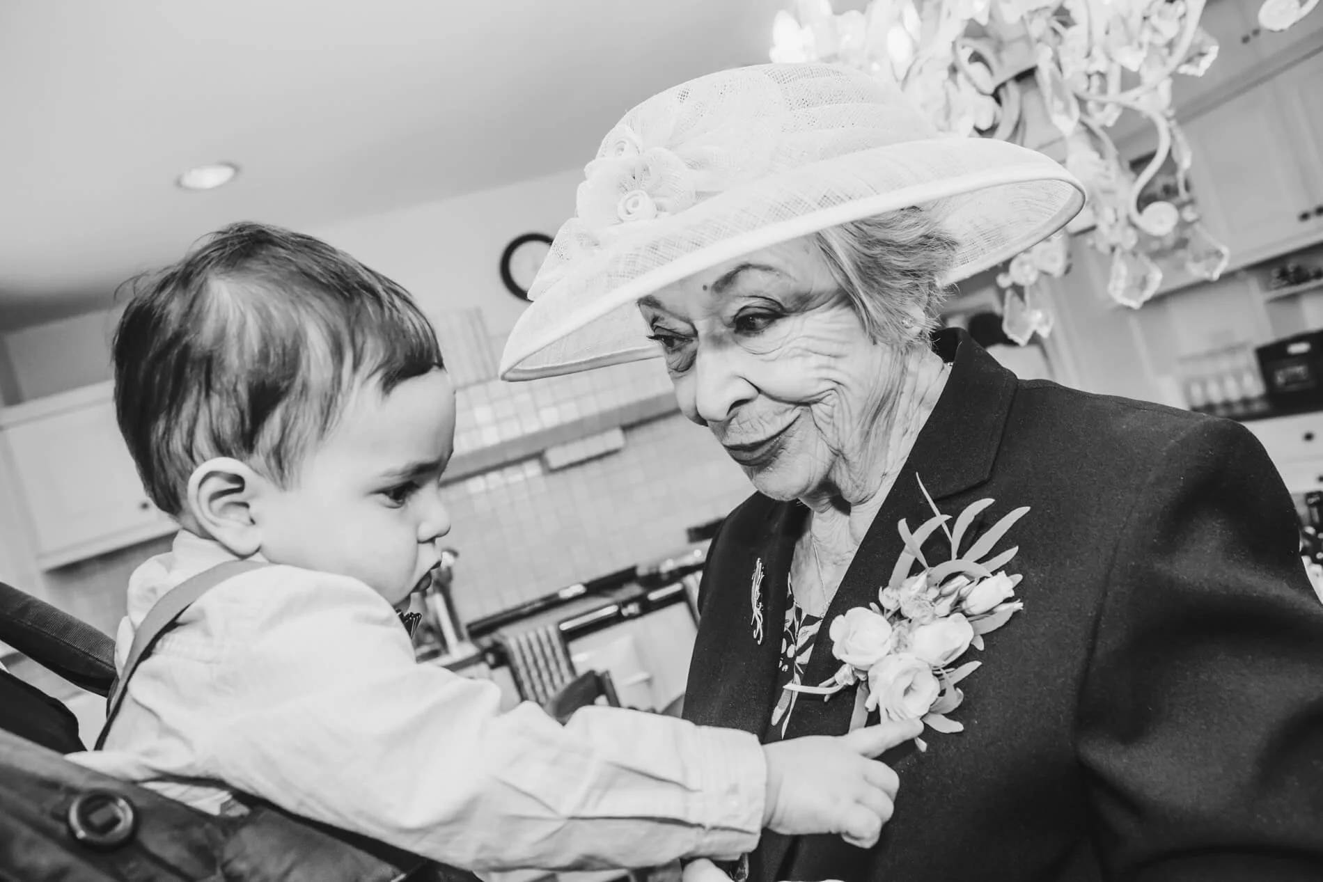 An elderly woman wearing a large hat and a dark coat with floral corsage, smiling as she interacts with a young boy in a stroller, pointing at her chest in the kitchen at Shortflatt Tower