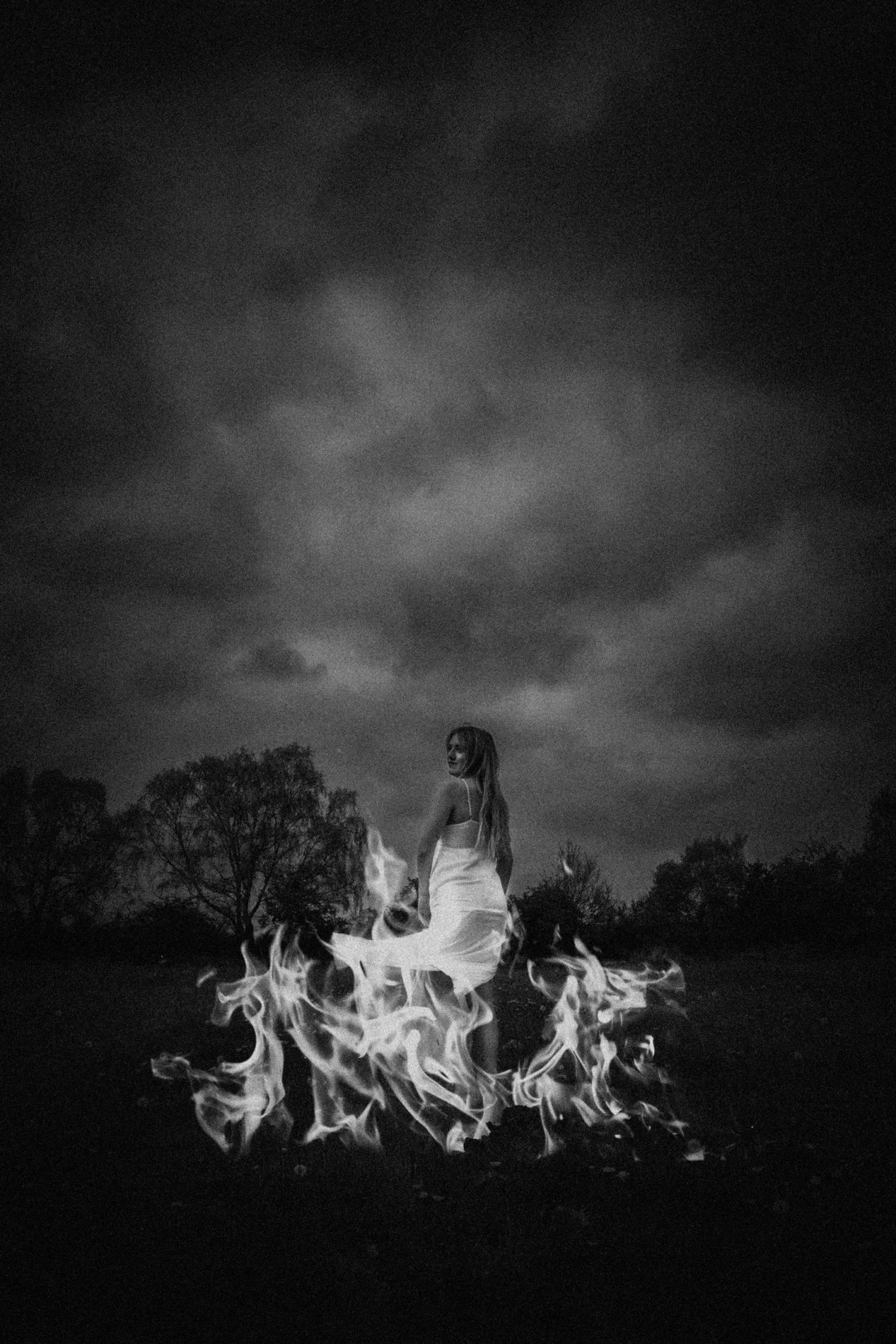 A black and white photo of a woman with long hair in a flowing white dress standing in the middle of flames on a grassy field with trees in the background, under a cloudy sky.