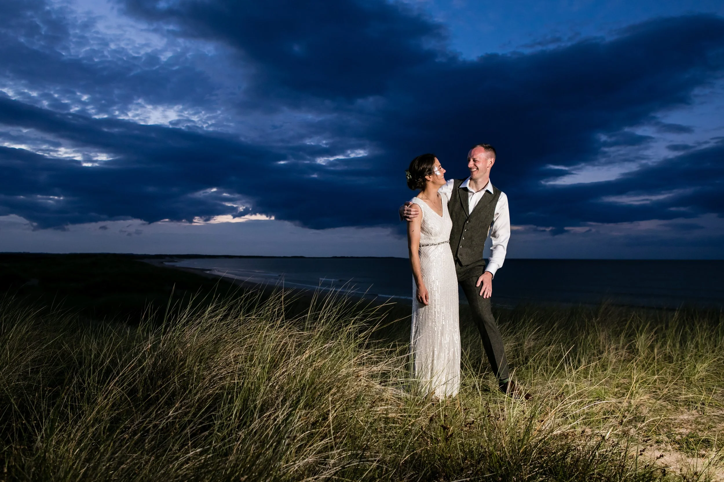 Newlyweds smiling at each other against a dramatic dark blue dusk sky on a grassy Northumberland dune, captured with soft, natural evening light.