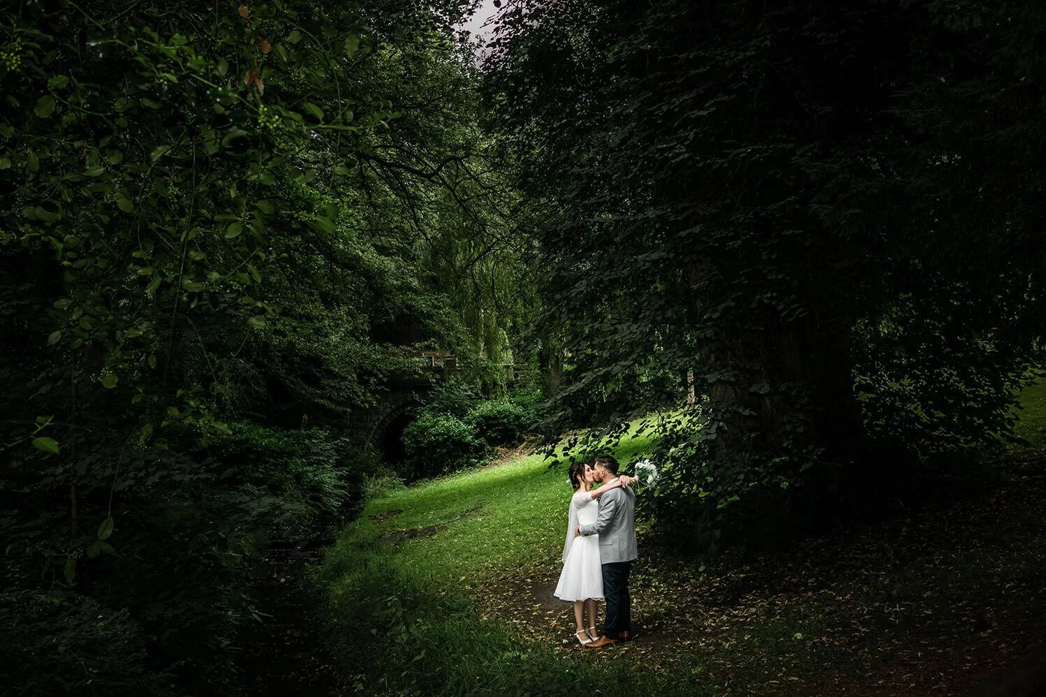 A couple dressed in wedding attire sharing a kiss in a lush green forest.
