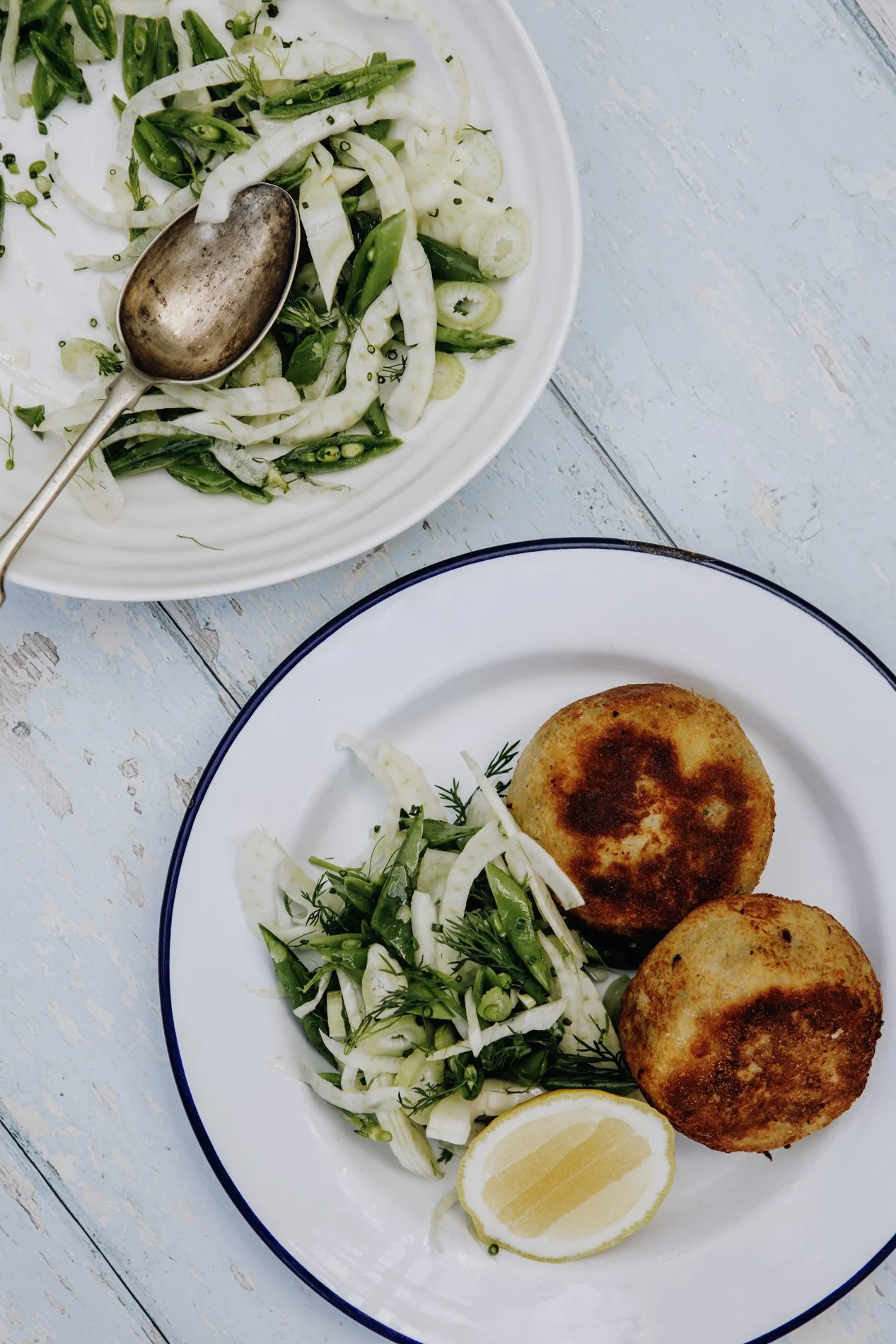 Plate with two golden brown potato croquettes, lemon wedge, and mixed salad with sliced fennel, peas, and herbs. Empty bowl of salad on a rustic light blue wooden surface.