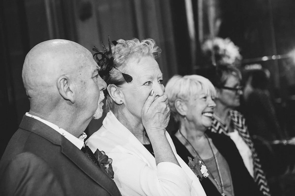 The bride's parents gasp as they see the bride for the first time. Their reactions are in close up, with the wedding officiants standing behind them smiling. This is in the great hall at Matfen Hall