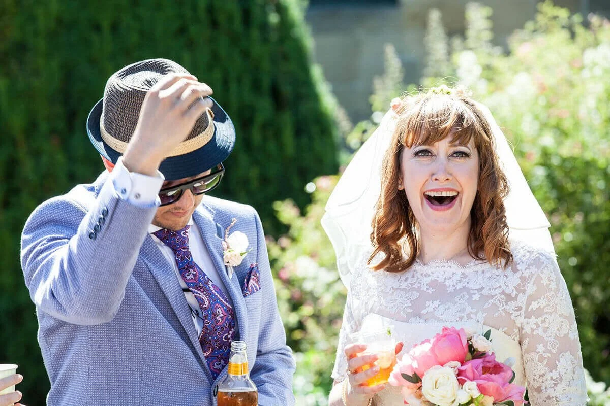 A woman in a wedding dress holding a bouquet of pink and white flowers, smiling happily; a man in a light blue suit, hat, and sunglasses adjusting his hat, standing outdoors on a sunny day with greenery in the background. 