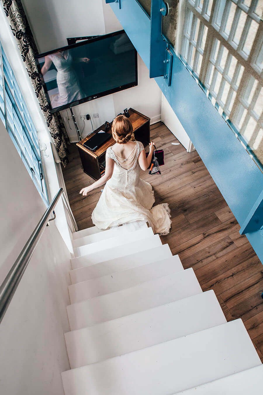 A woman in a wedding dress standing at the bottom of stairs, walking towards a television in a living room with wooden flooring and a glass block wall.