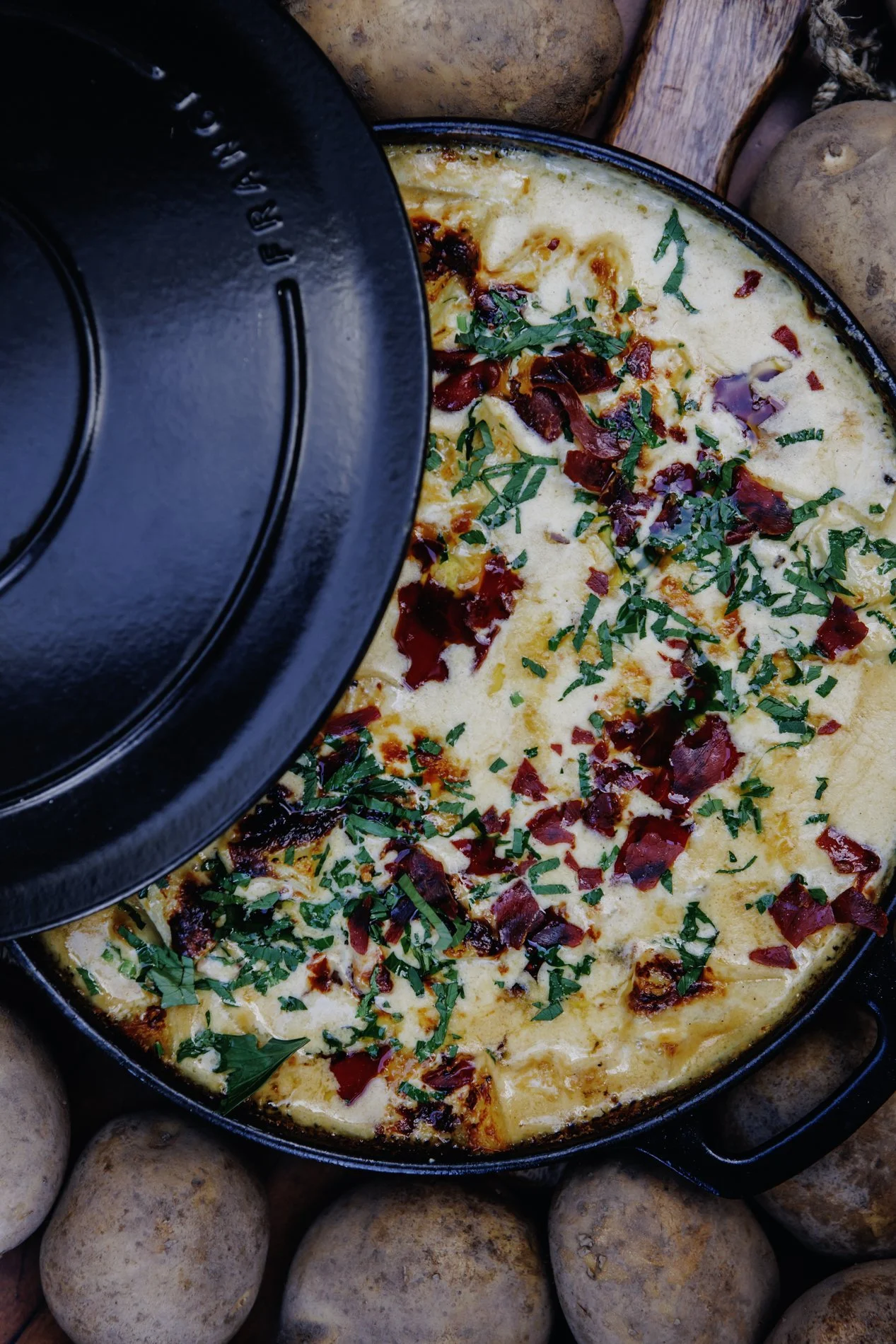 A skillet of cheesy breakfast casserole topped with chopped green herbs and bacon bits, surrounded by potatoes.