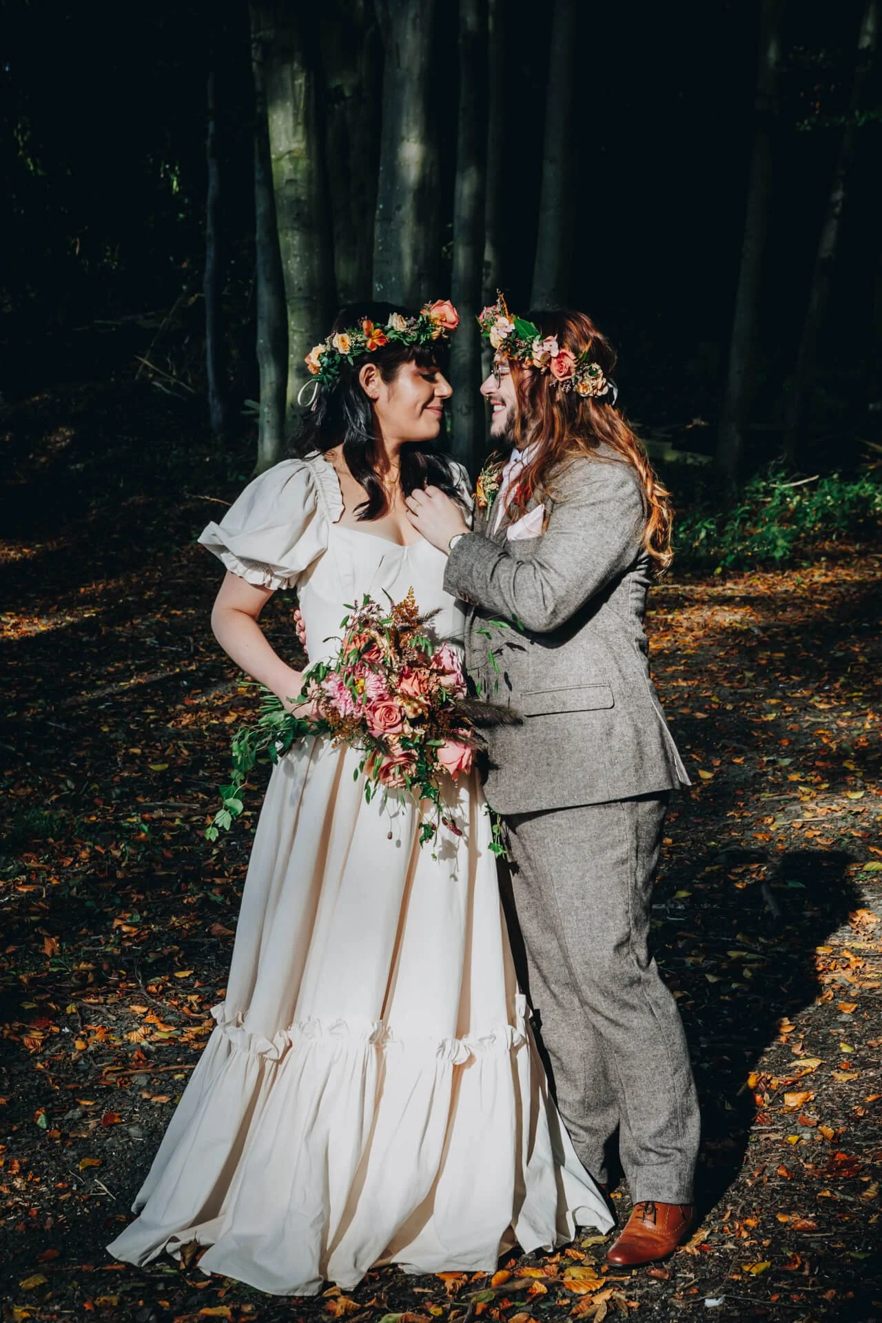 A couple in wedding attire embracing in a forest, wearing flower crowns and holding a bouquet of flowers.