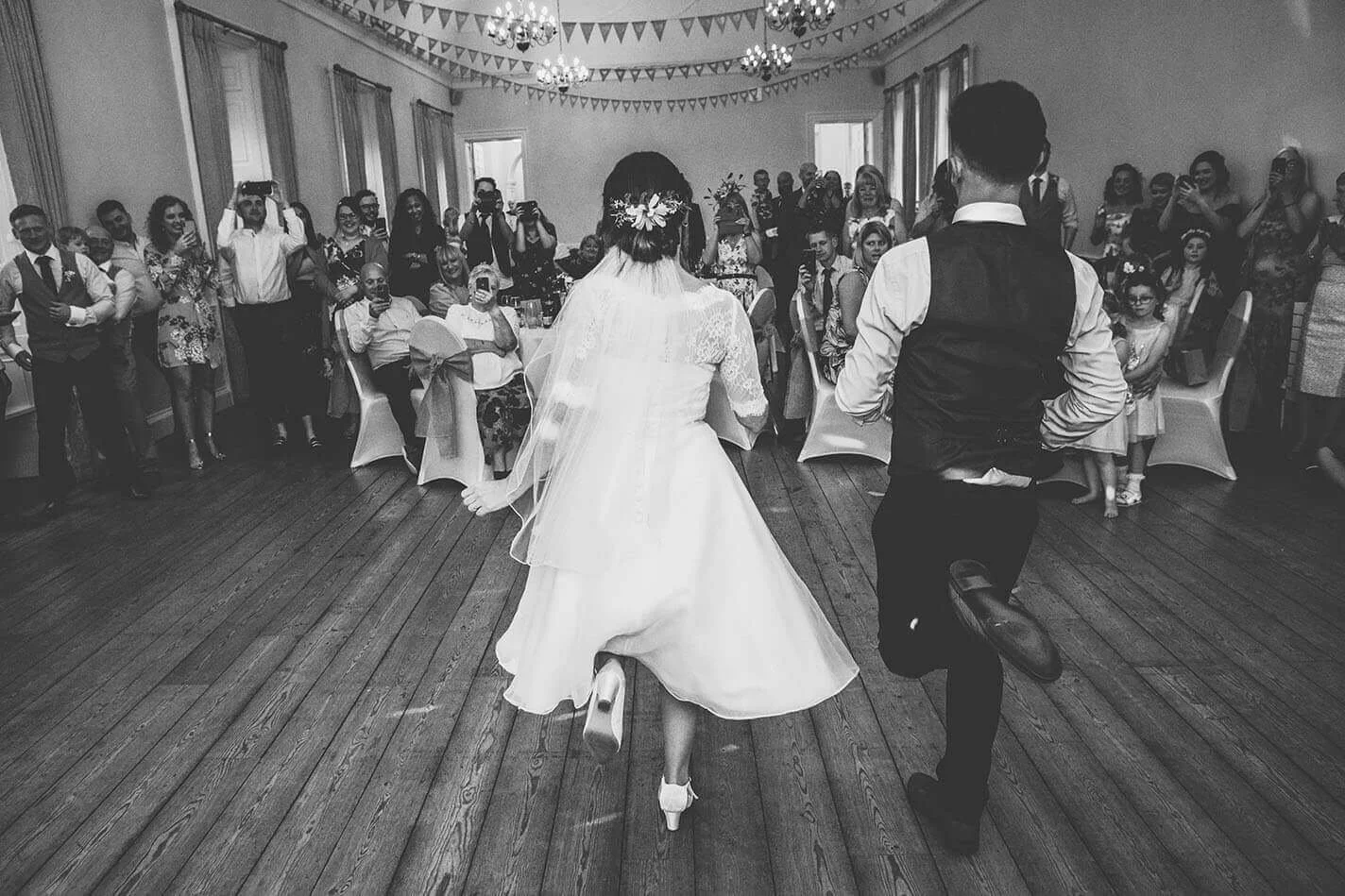 Black and white photo of a bride and groom dancing at a wedding reception, with guests watching and taking photos in a decorated hall at the top of Hexham Abbey