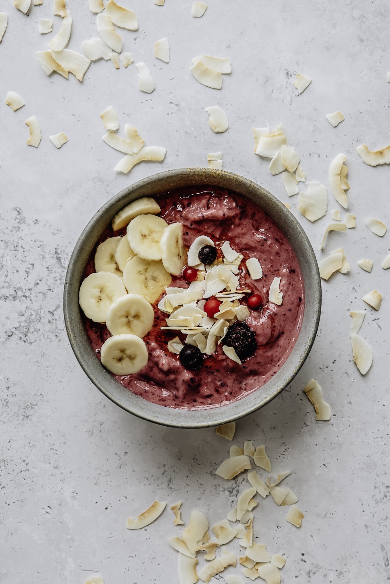 A bowl of berry smoothie topped with banana slices, white chocolate shavings, and mixed berries, on a white surface with scattered white chocolate flakes.