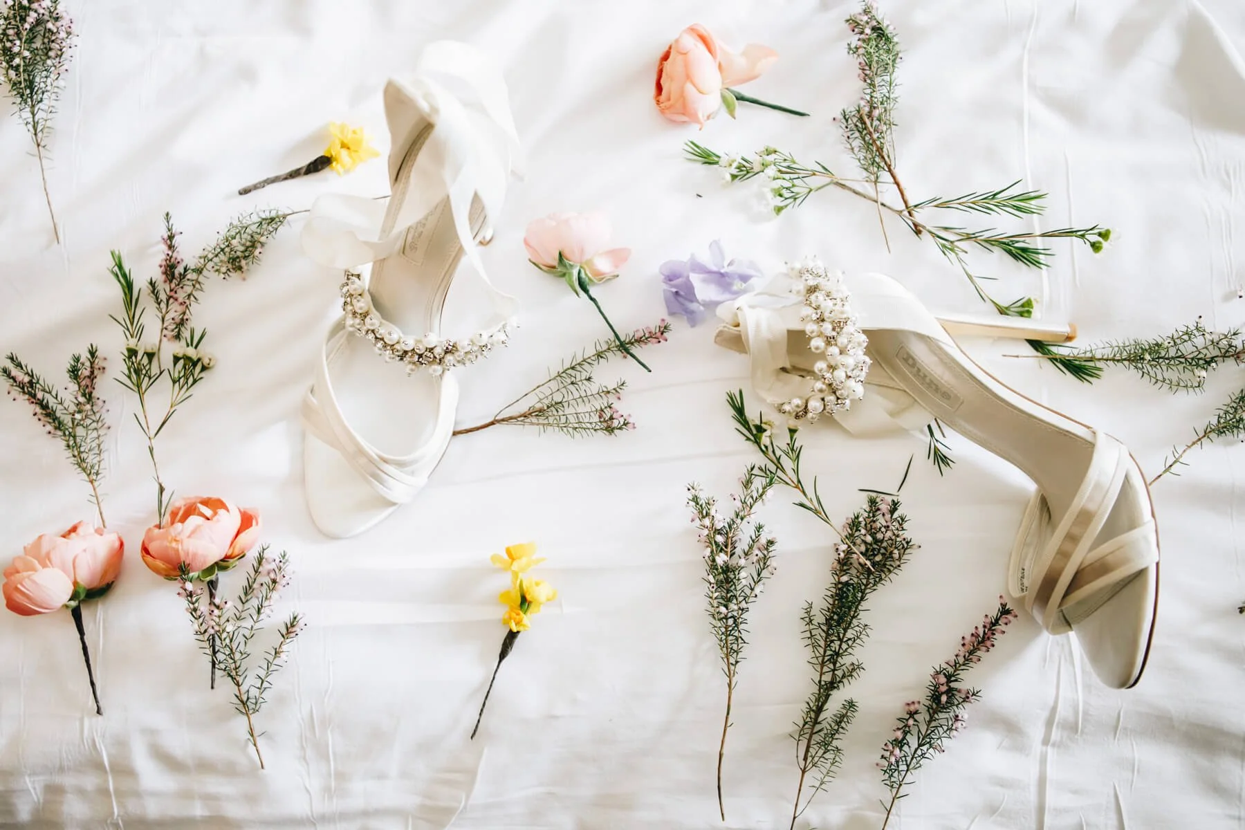 White satin bridal shoes with decorative pearl and rhinestone embellishments, surrounded by various pink, yellow, purple, and green flowers and foliage on a white fabric background. Image taken at Hotel du Vin
