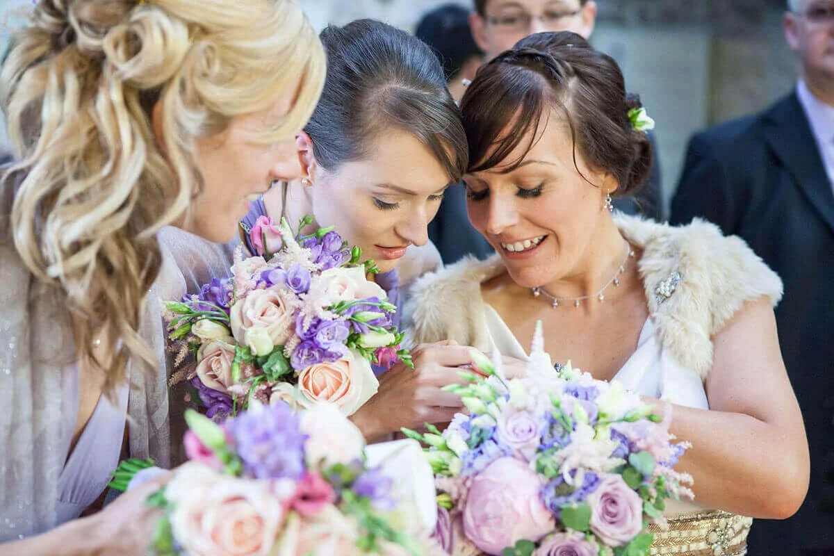 Three women smiling and holding bouquets of purple, pink, and white flowers at a wedding celebration. The bride is showing off her wedding ring at Matfen Brewery