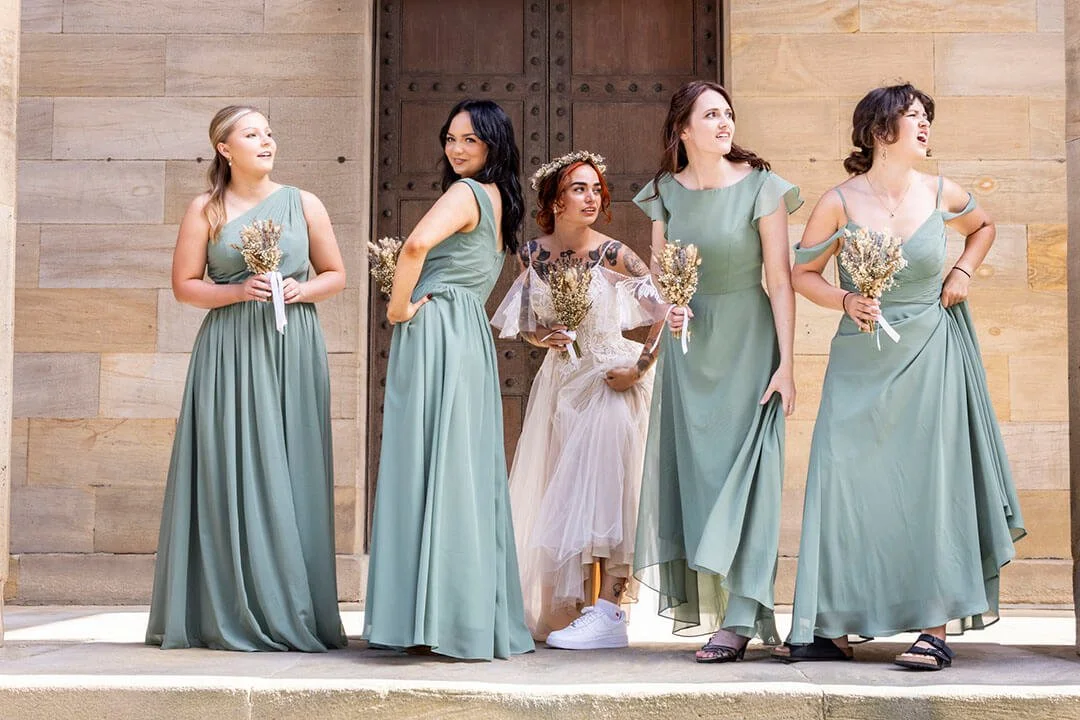 Five women in matching green dresses holding bouquets, standing in front of a wooden door, with one woman in a white dress with floral details, all appearing to be bridesmaids or at a wedding.