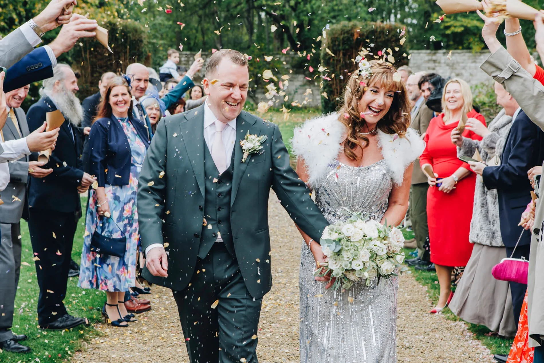 A newly married couple walking down a garden path at Shortflatt Tower, smiling, surrounded by guests throwing confetti at them at their wedding celebration.