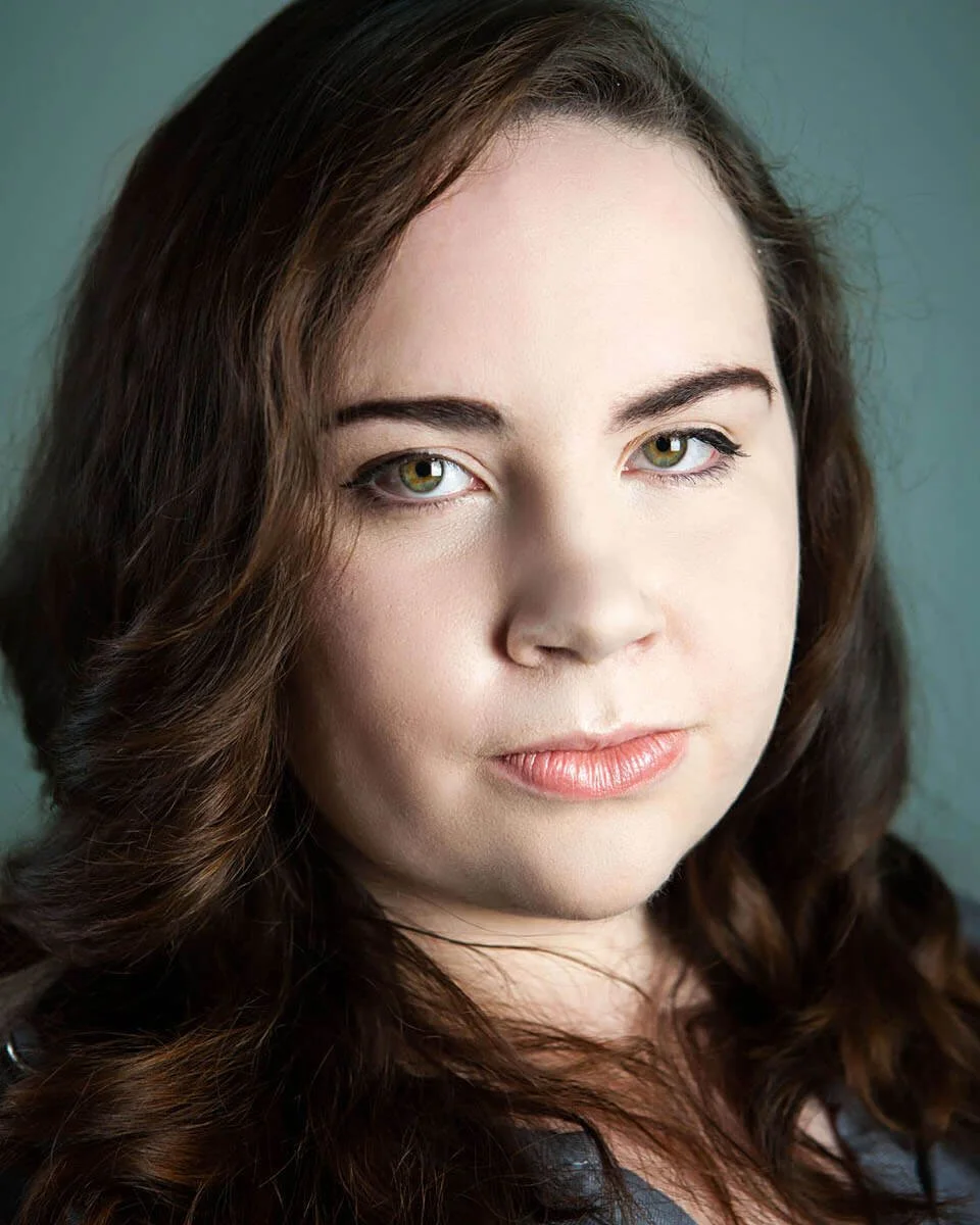 Close-up of a woman with wavy brown hair and green eyes, looking at the camera with a neutral expression.