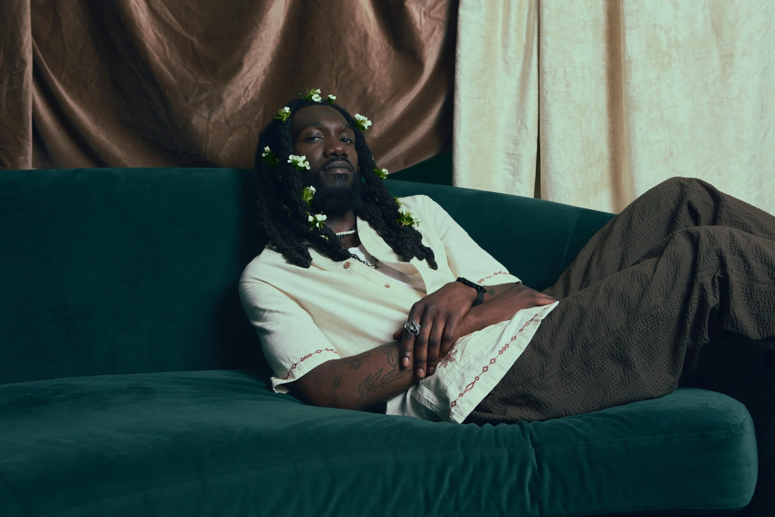 A man with dark skin, dreadlocks, and floral decorations on his head and beard, reclining on a dark green couch with crossed arms, wearing a white shirt with embroidery and brown pants, in a room with beige and pink velvet curtains.