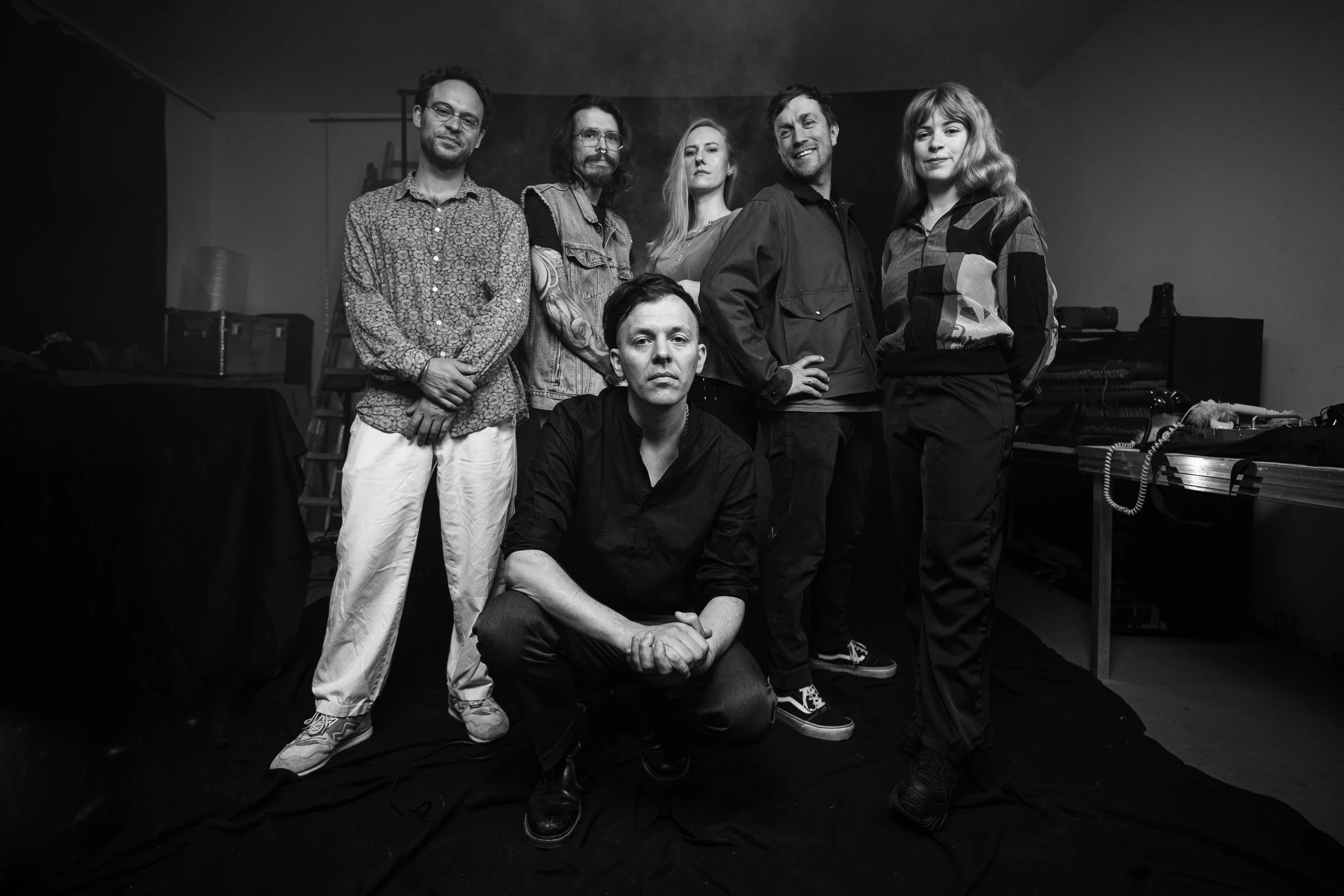 Group of seven people posing together indoors, some standing and one kneeling, with musical equipment and a piano in the background.