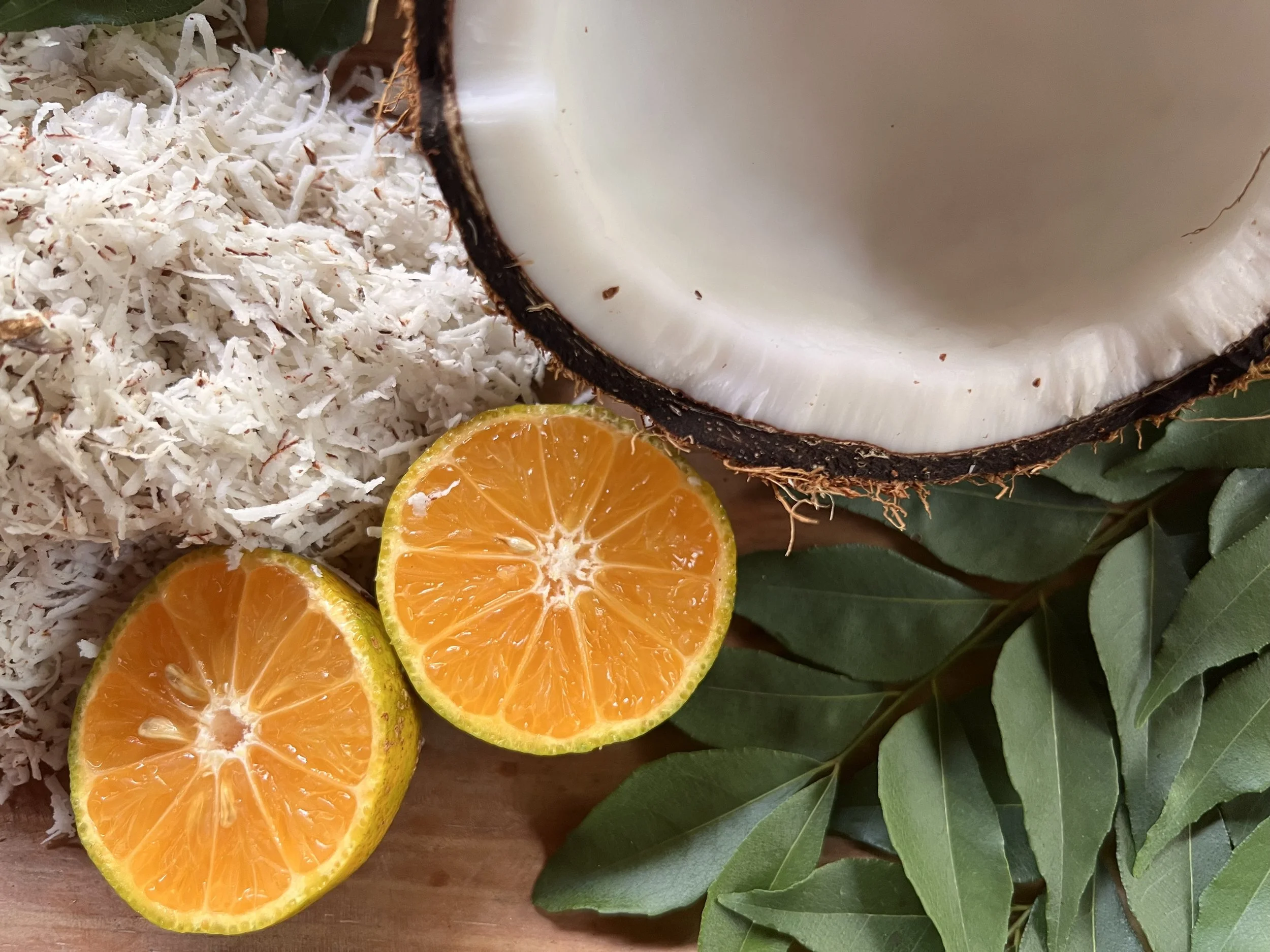 Half a coconut with white flesh, two halved oranges showing their bright orange interior, shredded white coconut, and green leaves.
