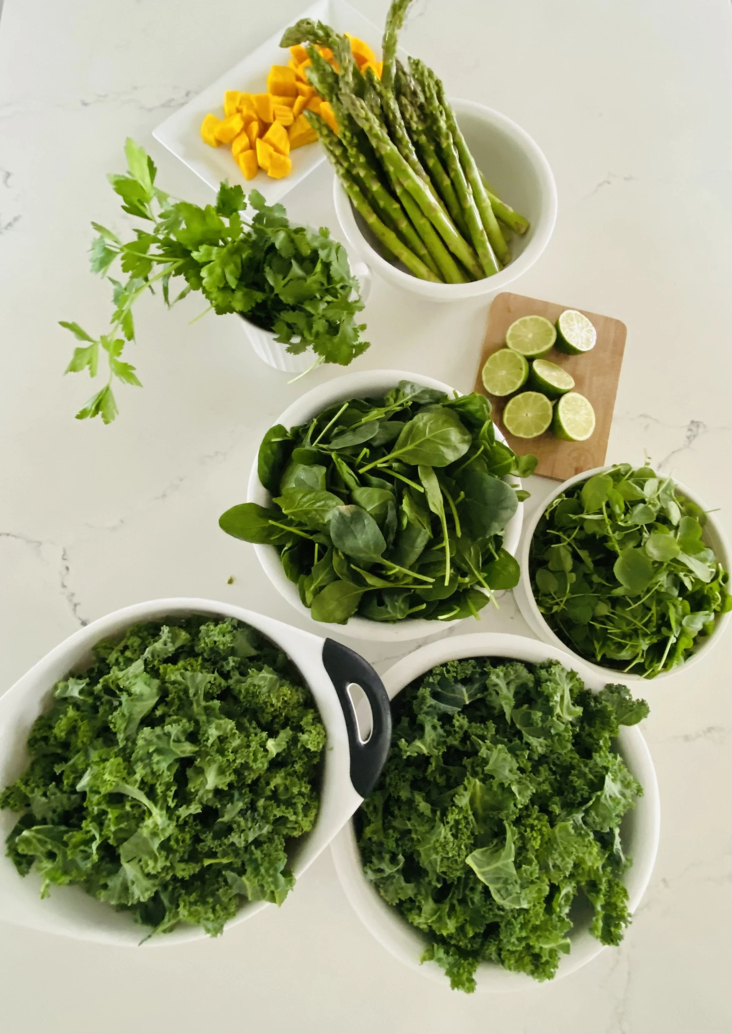 assortment of fresh vegetables and herbs including kale, spinach, microgreens, cilantro, asparagus, lime, chopped mango, and other greens on a white surface.
