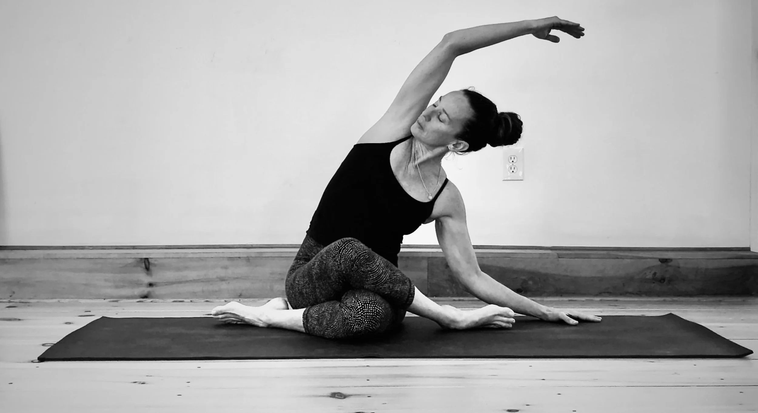 A woman performing a seated side stretch yoga pose on a black yoga mat in a room with wooden floor and plain wall.