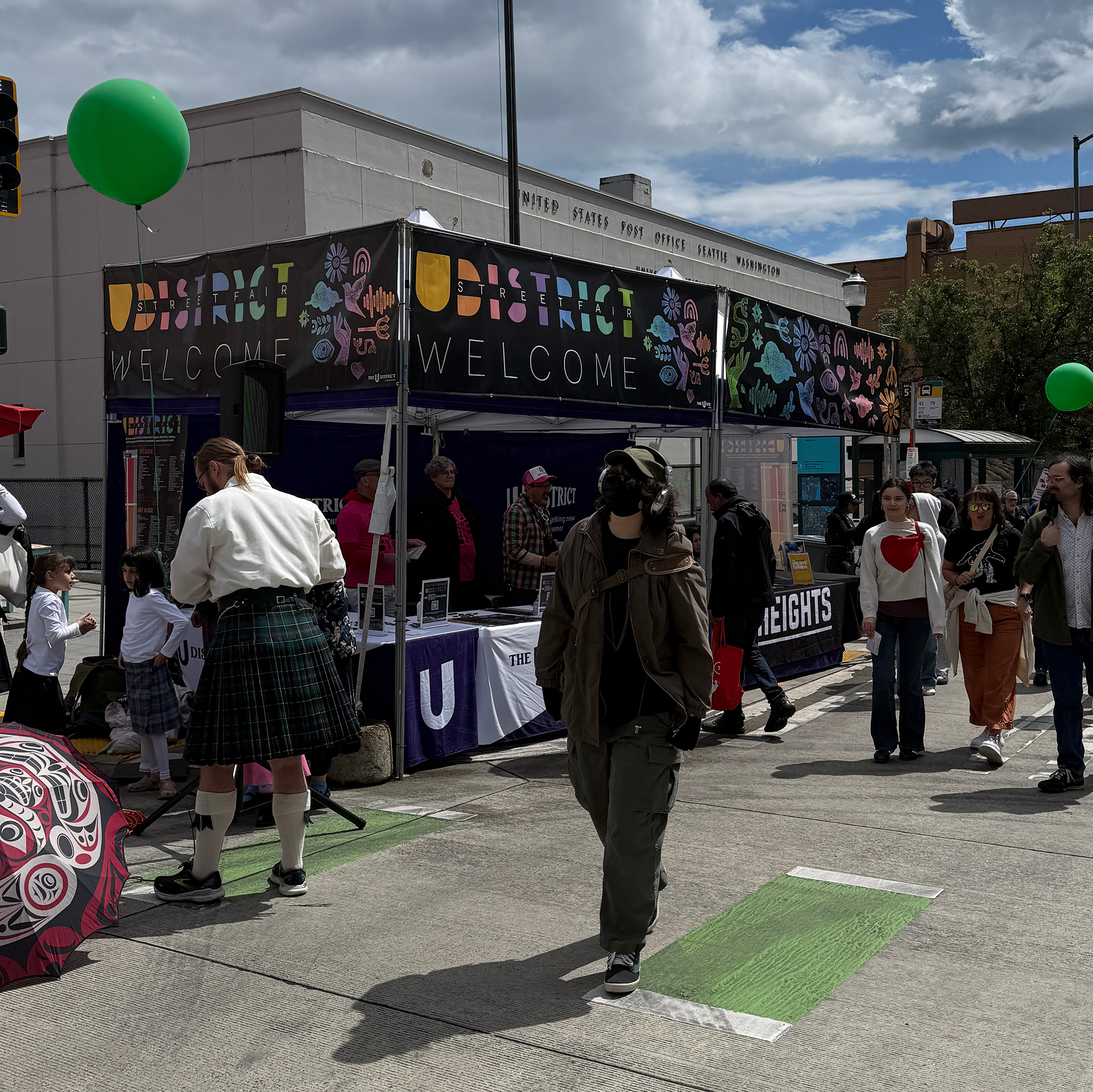Street festival scene outside the United States Post Office in Seattle, Washington. People gather around a tent with a colorful banner that says 'District Street Fair' and 'Welcome.' There are green balloons and umbrellas, and the sky is partly cloud