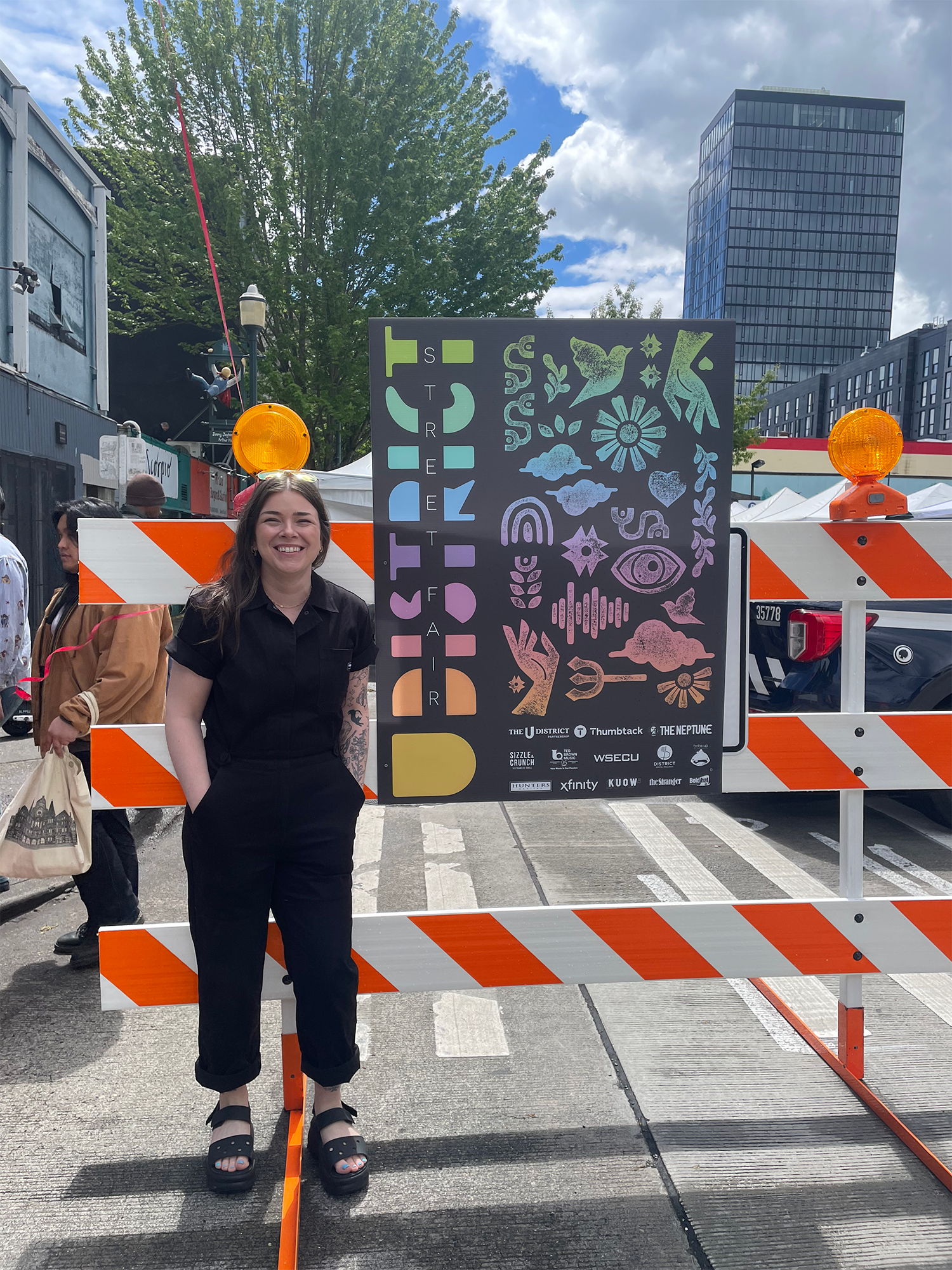 A smiling woman standing behind orange and white striped barriers at an outdoor event. Behind her, a large colorful sign with various symbols and the words "Streat" and "Taste" visible. Several tents and a building with glass windows can be seen unde