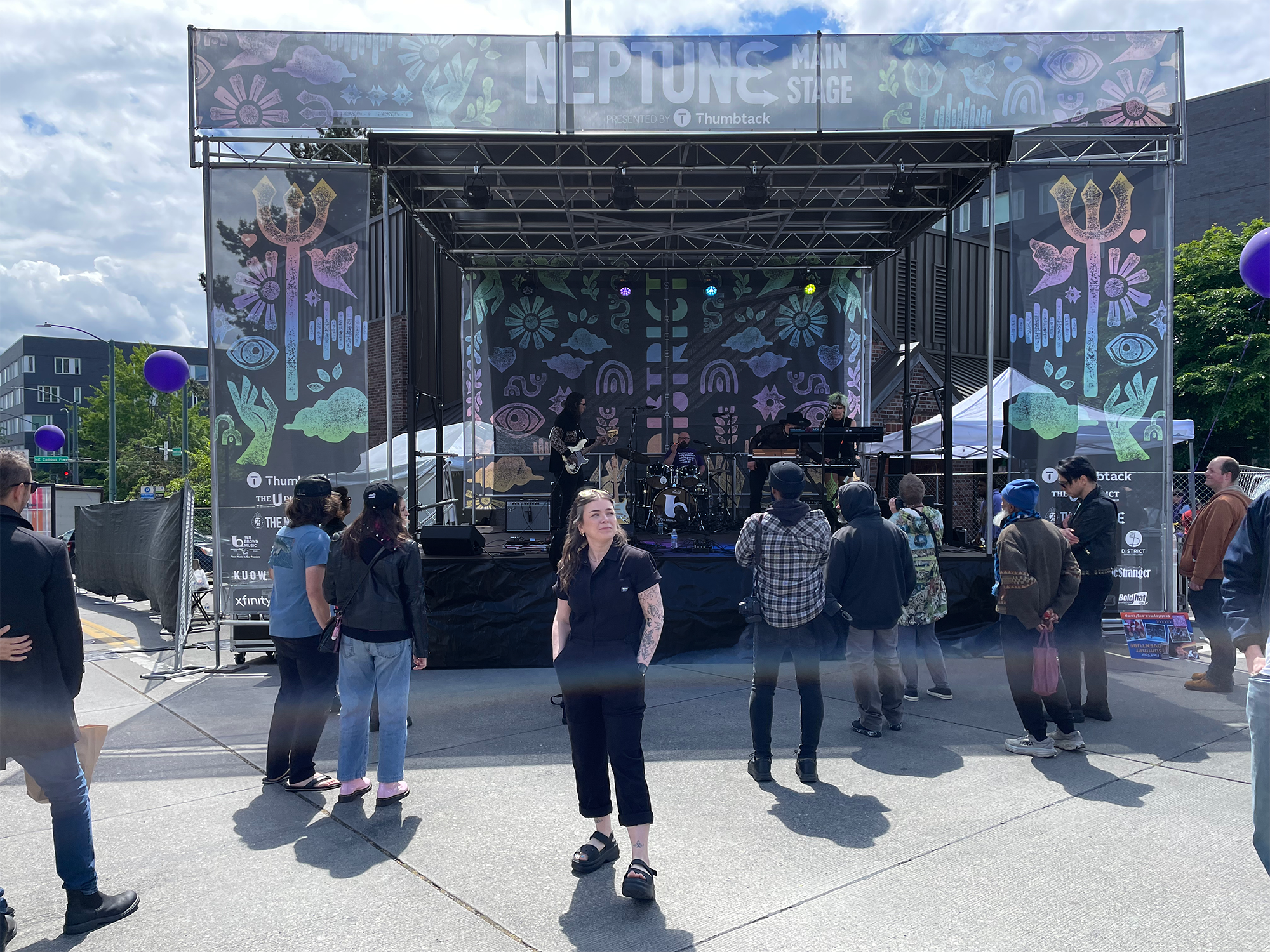 People attending an outdoor concert with a band performing on stage decorated with colorful abstract designs and symbols, under partly cloudy skies.