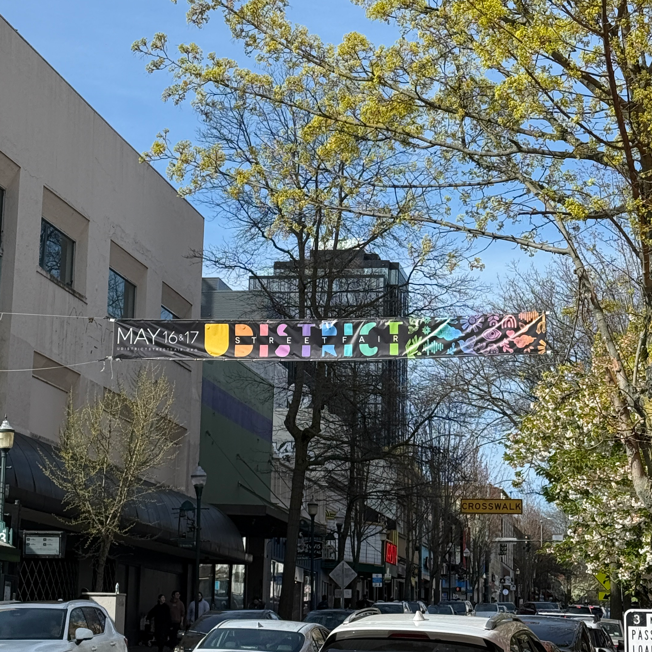 City street scene with cars parked along the street, pedestrians walking on the sidewalk, multiple commercial buildings, trees with budding leaves, and signs indicating a street fair event from May 16-17, with a crosswalk sign present.
