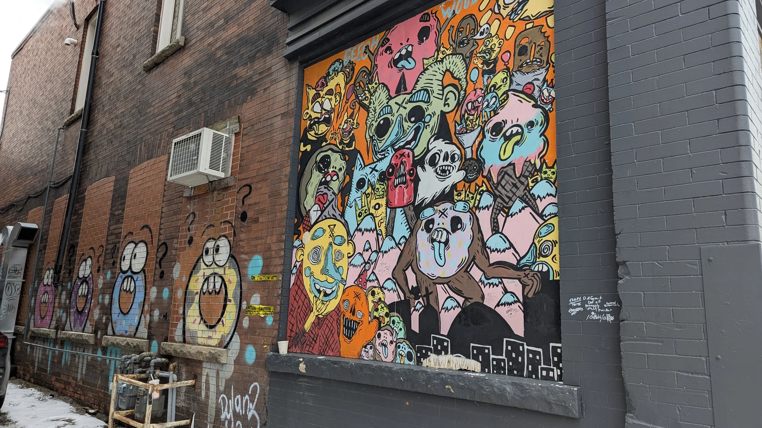 Colorful street art mural on a brick wall in St. Catharines featuring various cartoonish monster characters with exaggerated facial expressions, some holding ice cream cones and others with their tongues sticking out.