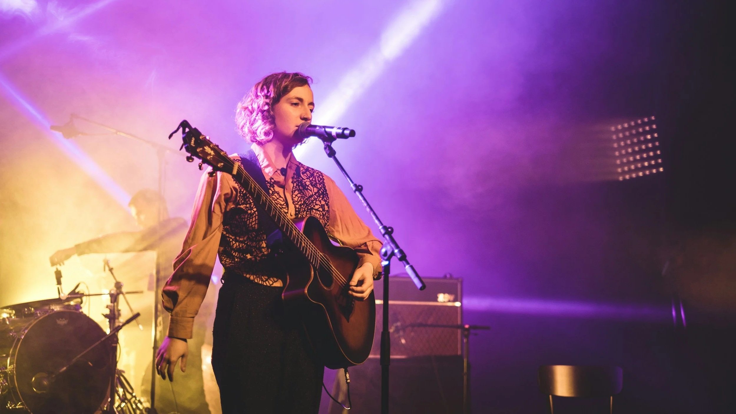 A woman with short curly hair singing into a microphone while playing an acoustic guitar on stage with colorful purple and yellow lighting.