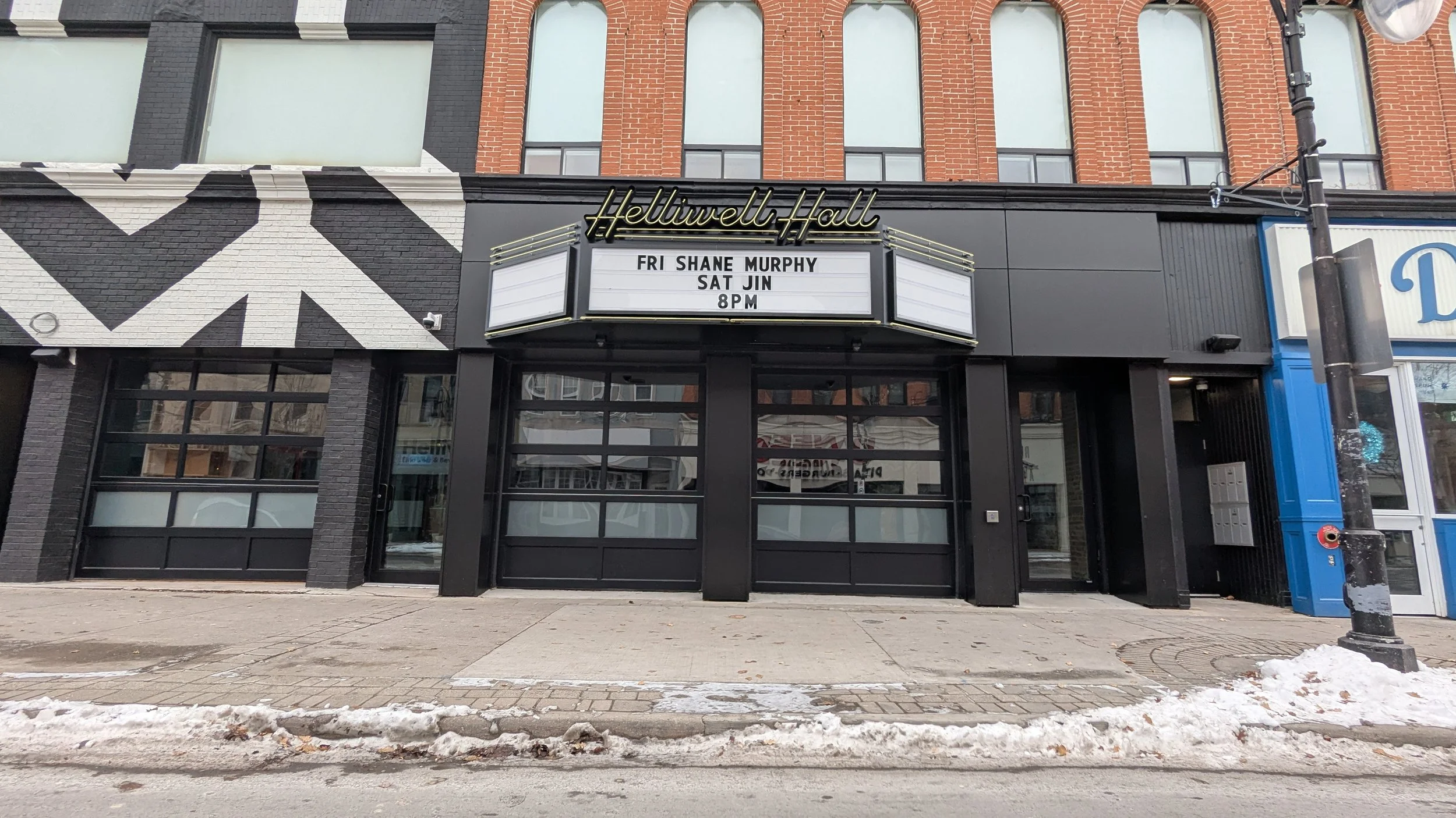 The exterior of Helliwell Hall theater in St. Catharines with a marquee sign displaying upcoming show with details, set on a city sidewalk with snow patches.