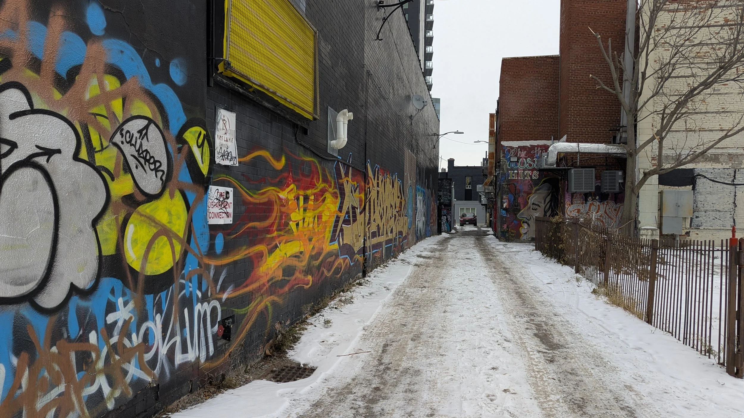 A snowy alleyway with colorful graffiti on the walls in St. Catharines, and a few signs posted on the left wall. A bare tree is on the right side near a fence.