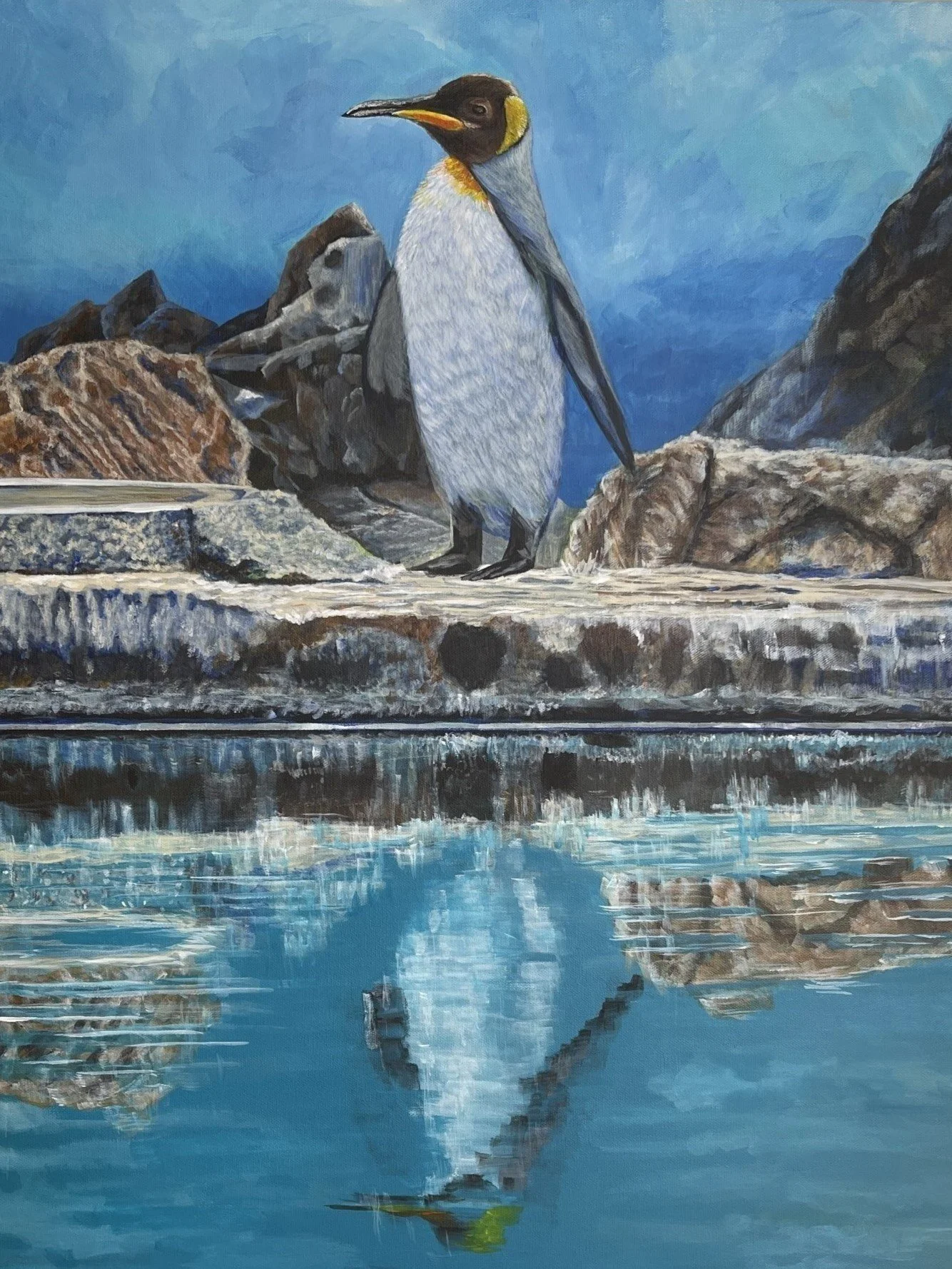 Painting of a penguin standing on rocky terrain near water with a reflection of the penguin and rocks in the water.