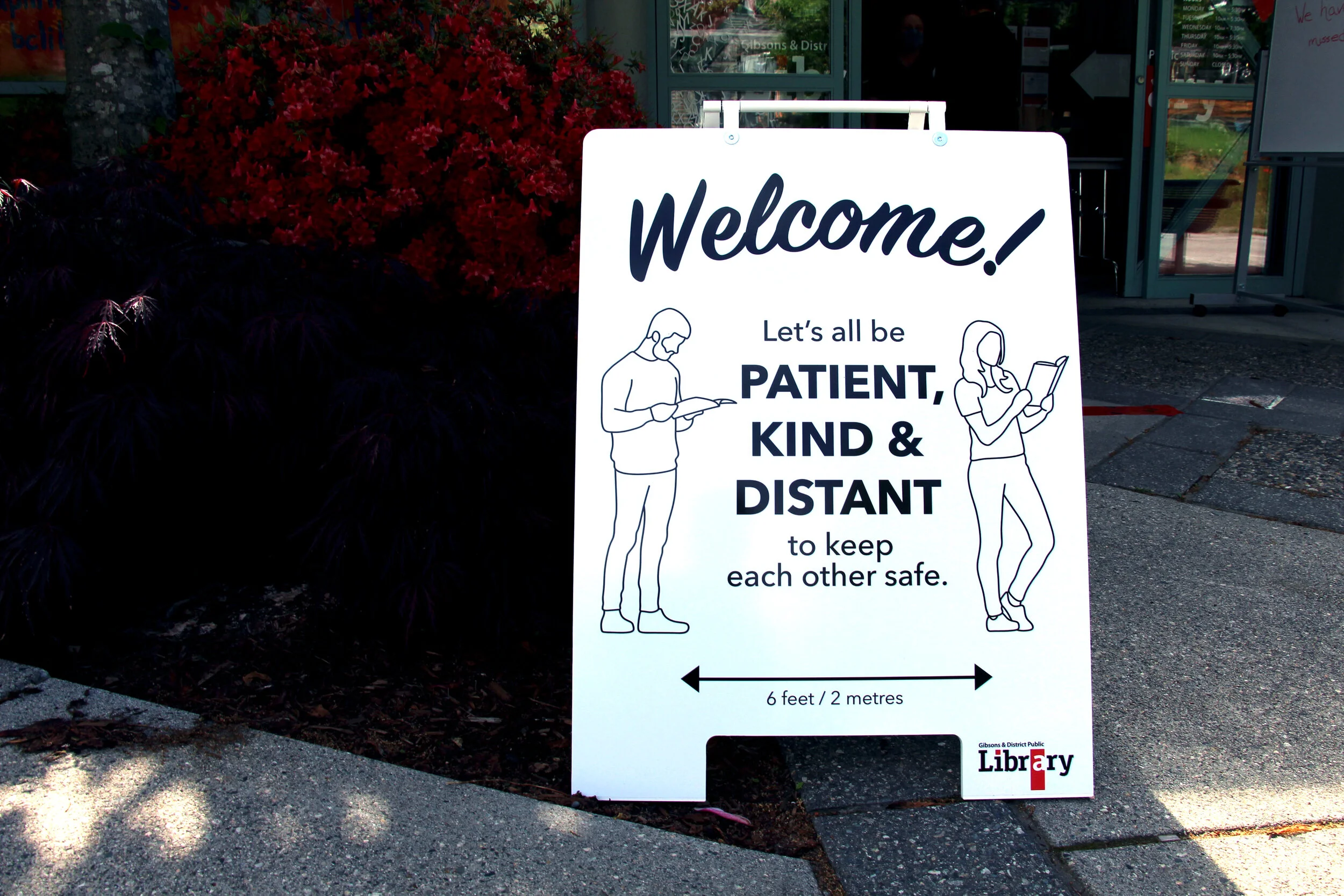 white sandwich board in front of a library with the line drawing of two people seperated by the following text "Weclome! Let's all be patient, kind & distant to keep each other safe. (6 feet/ 2 meters)"