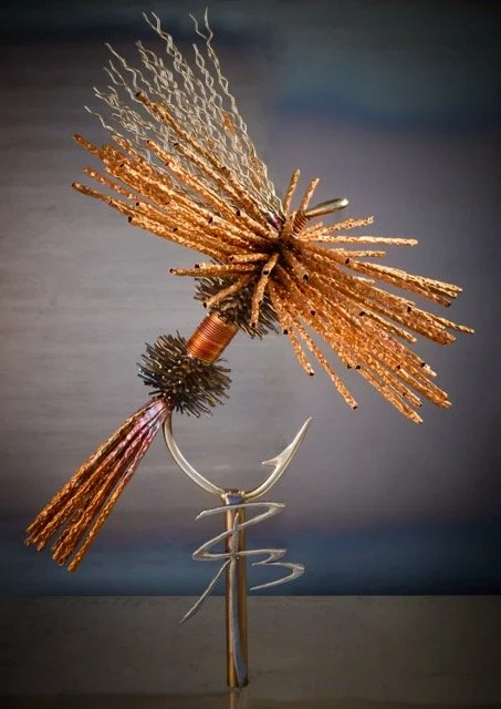 Colorful metal sculpture of a bird with blue and orange feathers, mounted on a circular stand, against a black background.
