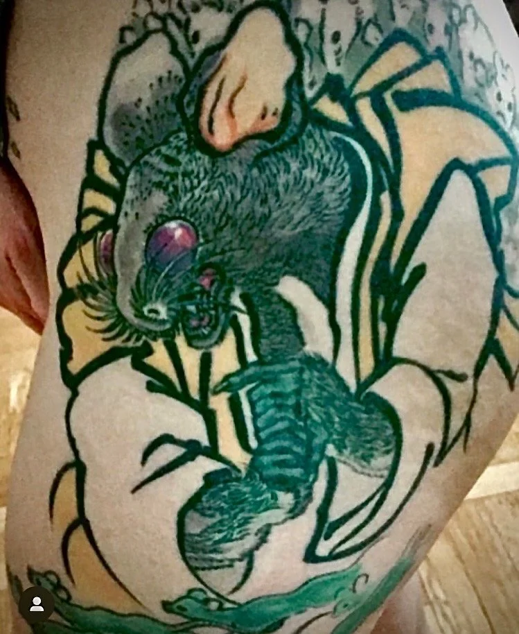 Tattoo of a rat with a red nose, surrounded by leaves and plant elements on skin.