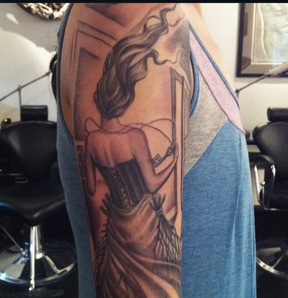 Tattoo of a woman with long hair seen from behind, sitting in a therapy or meditation room, with her back exposed, wearing a corset, holding a chain connected to a device or monitor.