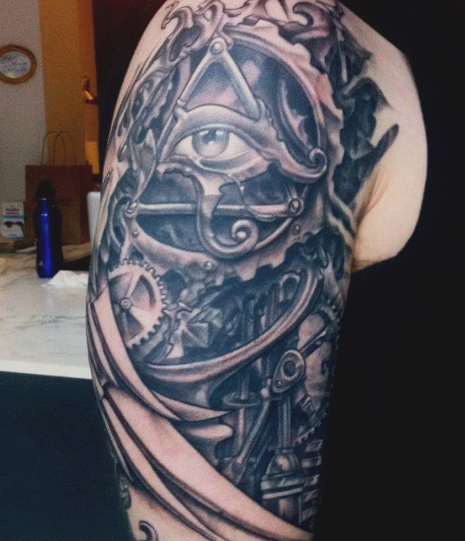 Black and gray tattoo of clockwork gears, chains, and a prominent eye on a person's upper arm and shoulder.