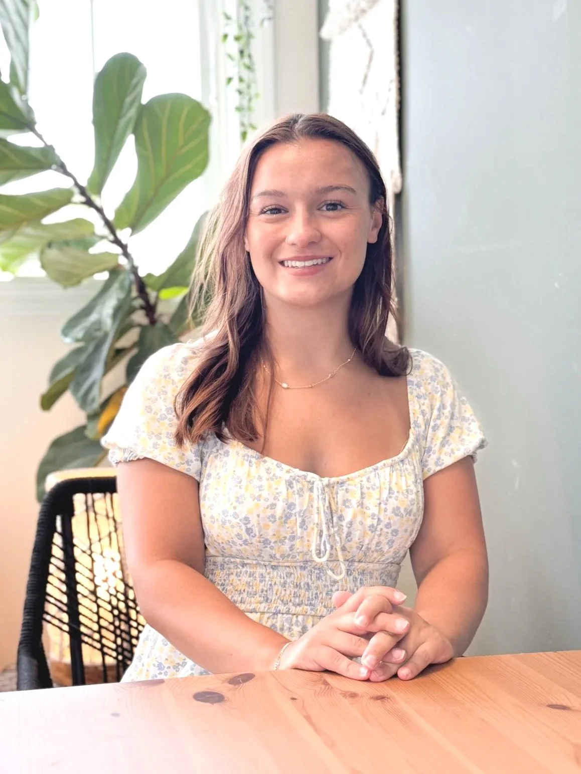 A young woman with brown hair and a floral yellow dress, sitting at a wooden table, smiling, with a green plant and window in the background.