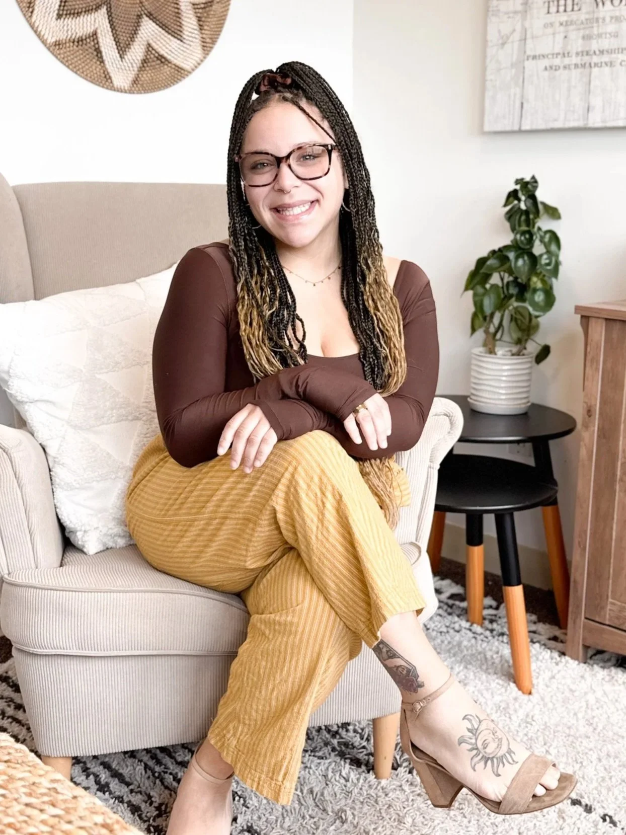 A woman with braids and glasses sitting on a beige couch, smiling. She's wearing brown top, yellow pants, and tan high heels with tattoos on her foot. Behind her is a small black table with a green potted plant, and there is artwork on the wall.
