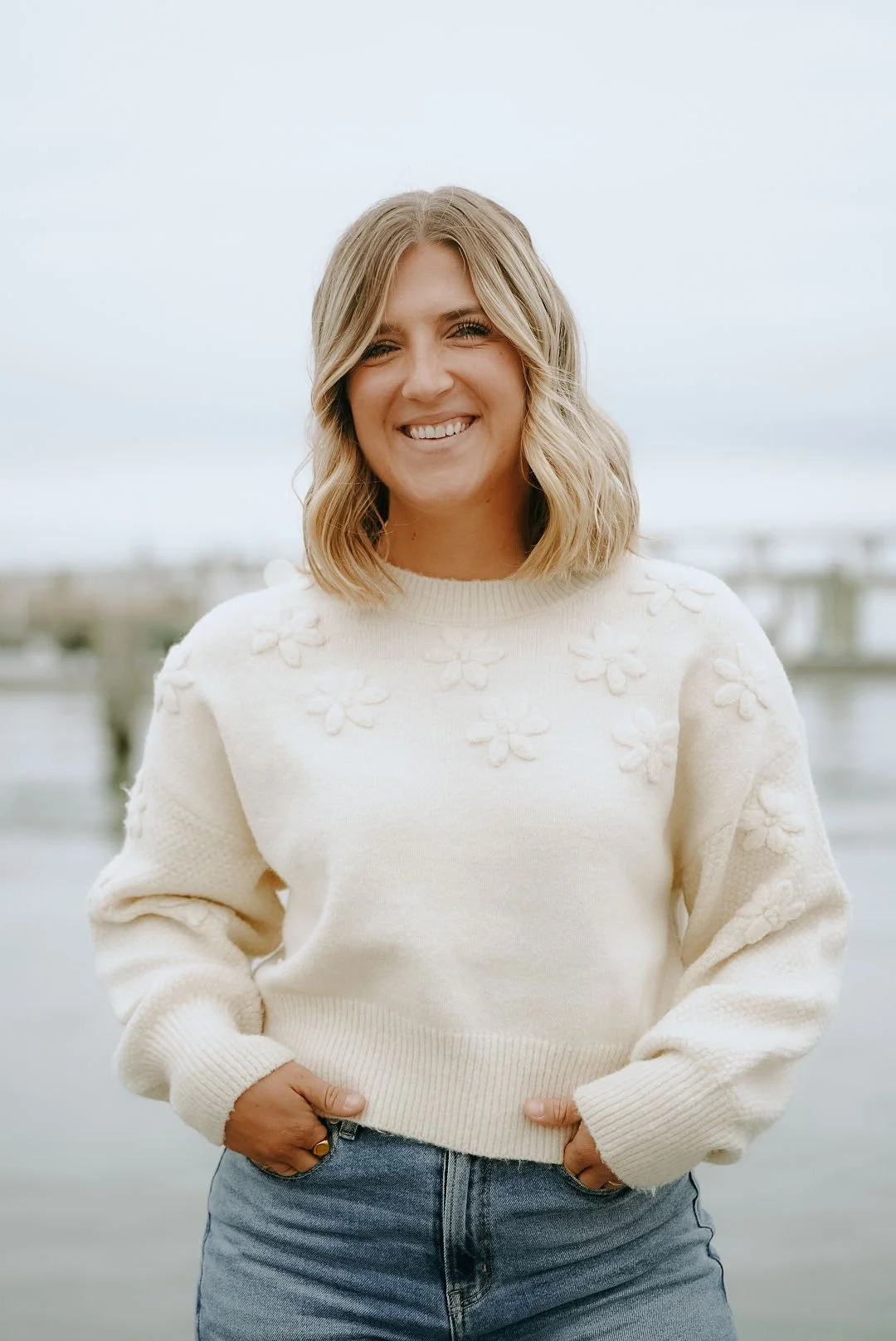 Young woman with blonde hair smiling, wearing a cream sweater with floral embroidery, standing outdoors near water with cloudy sky background.