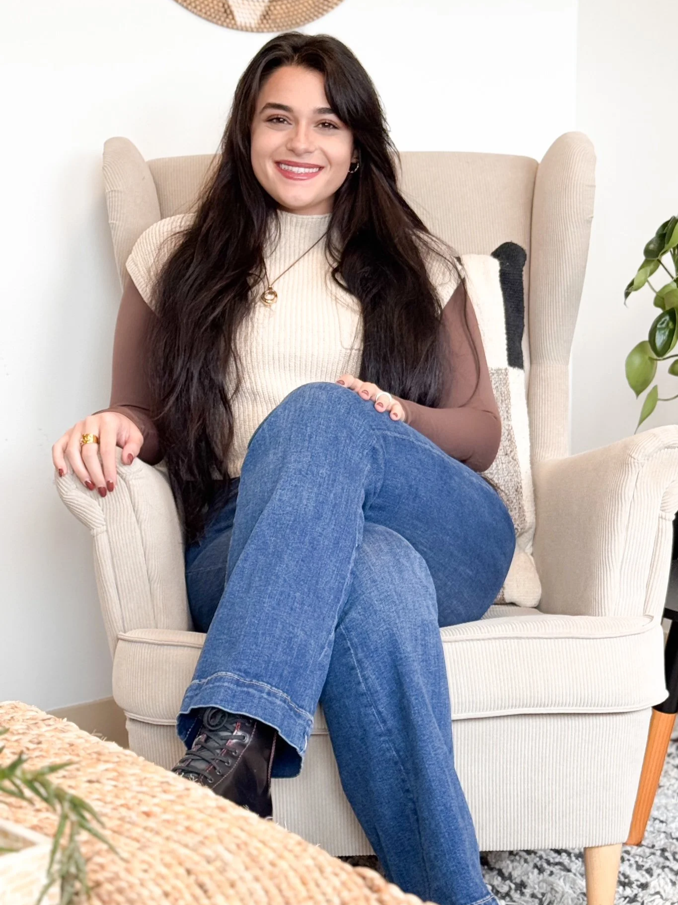 A woman with long dark hair sitting comfortably in a beige armchair, smiling, in a cozy room with a potted plant beside her.