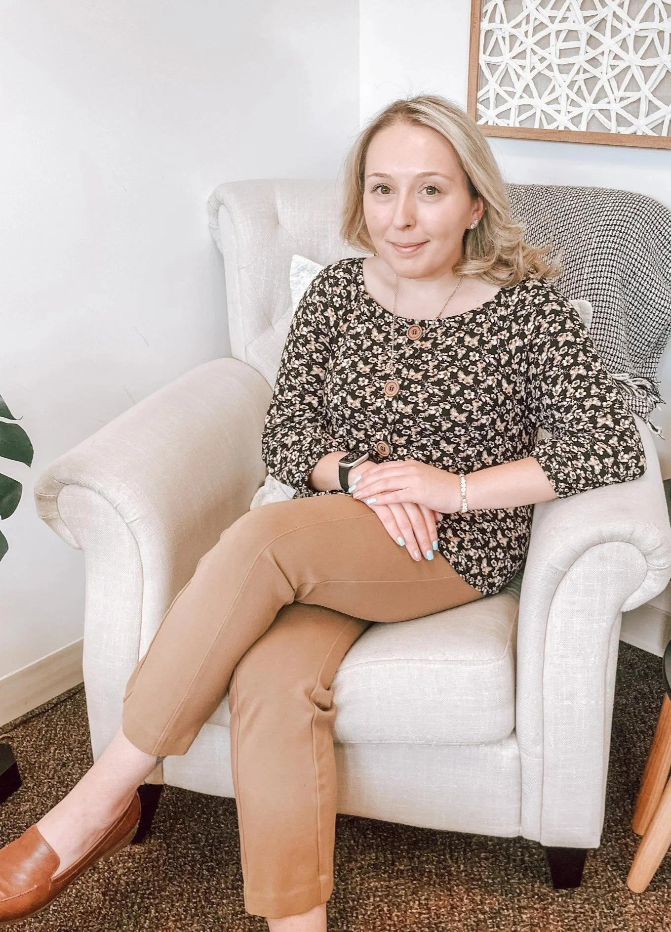 A woman with short blonde hair sitting in a beige armchair, wearing a floral blouse, tan pants, and brown shoes, smiling with a watch on her wrist and a bracelet, in a room with white walls and a decorative wall art piece.