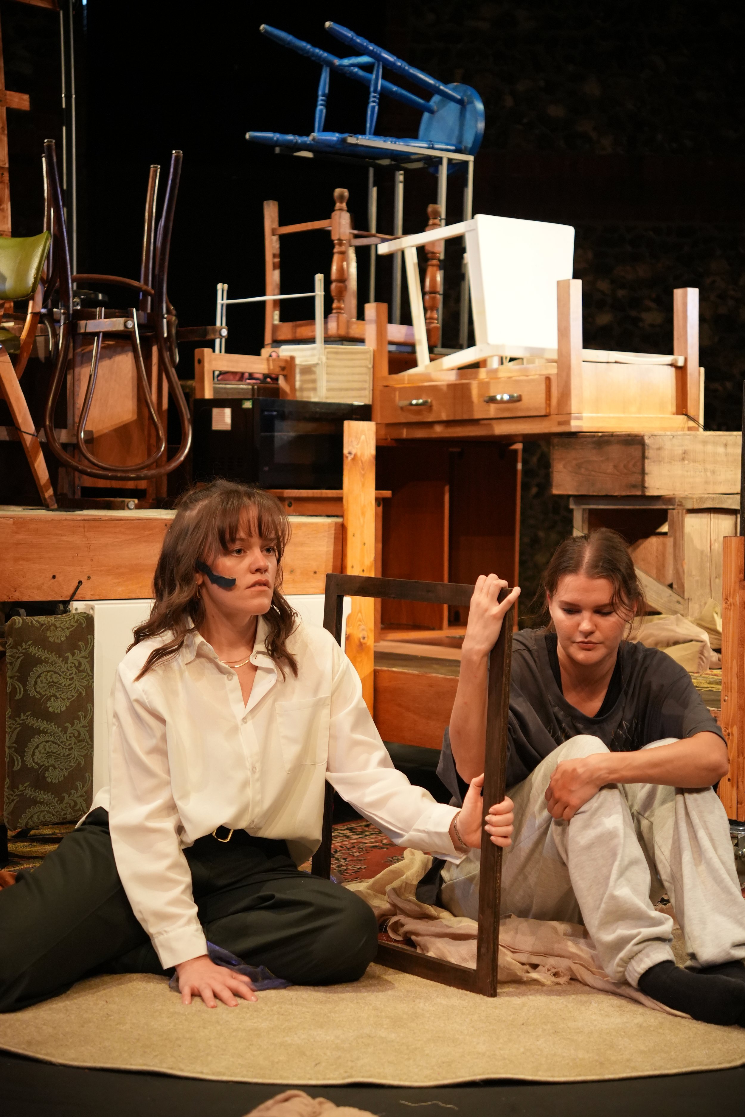 Two women sitting on the floor holding a frame between them as if it were a door. Taylor (left) has a scar on her face and looks desperately into the distance.