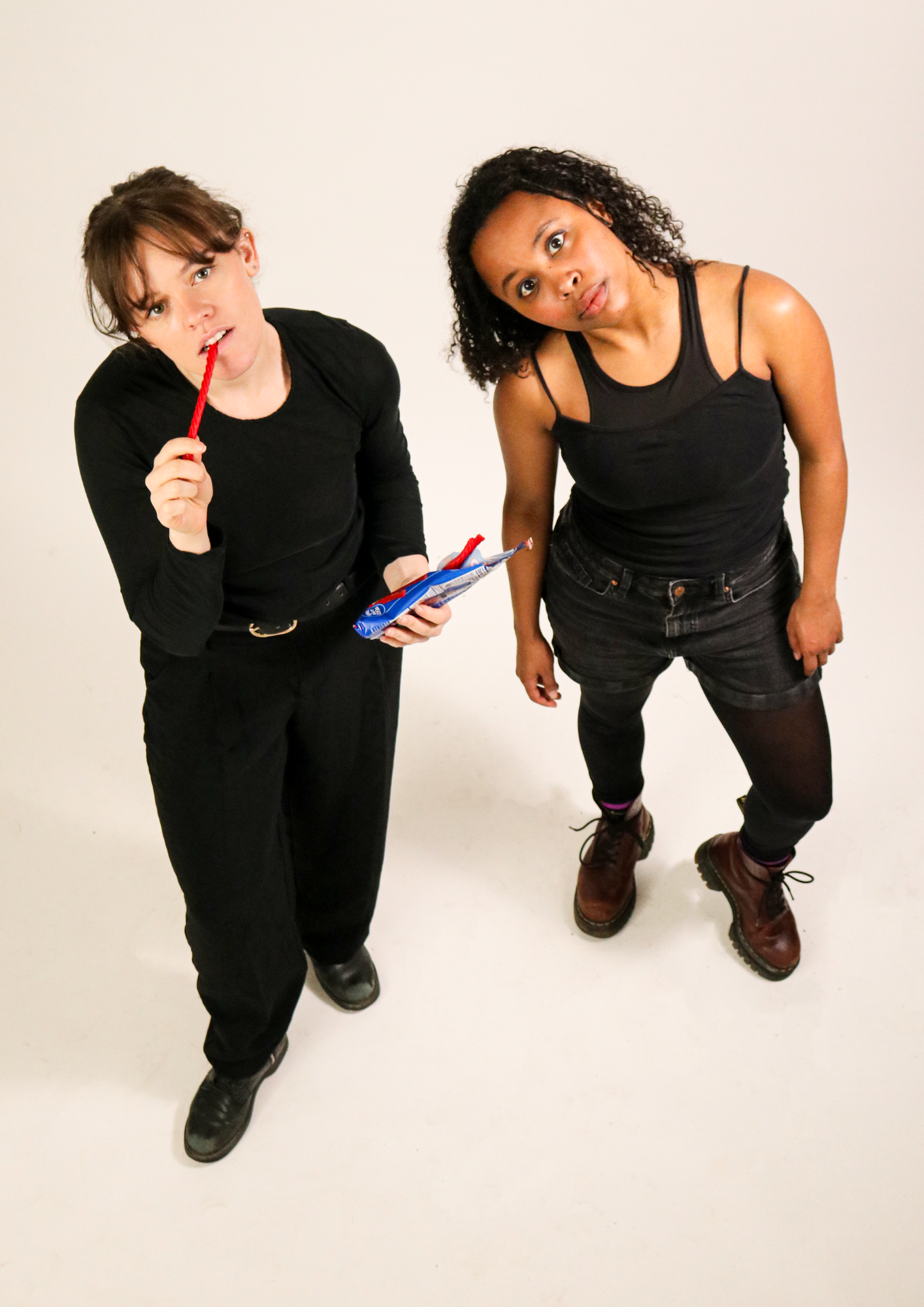 Two women standing on a white background staring up at the camera, eating a bag of candy.