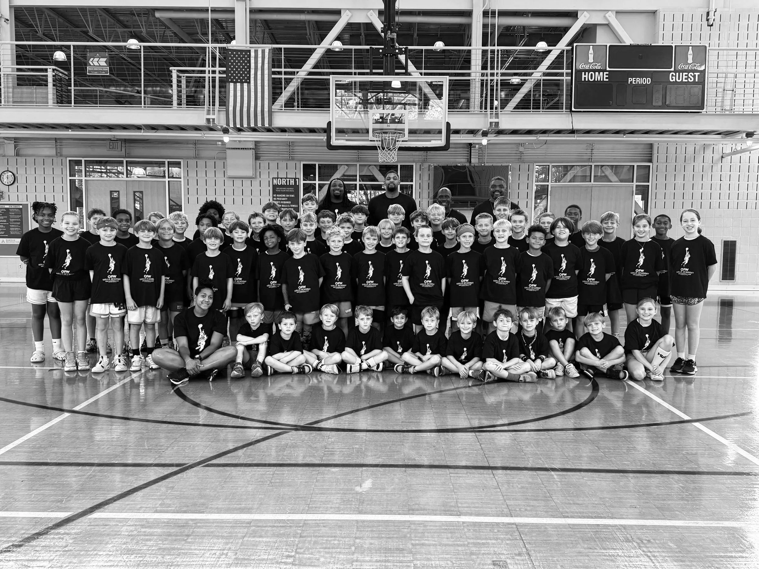 Group of children and their coaches posing for a photo on a basketball court inside a gymnasium. The kids are wearing matching T-shirts with a logo, and some are standing while others are sitting cross-legged in front. The gym has a basketball hoop, scoreboard, and American flag, with high ceiling and metal beams.