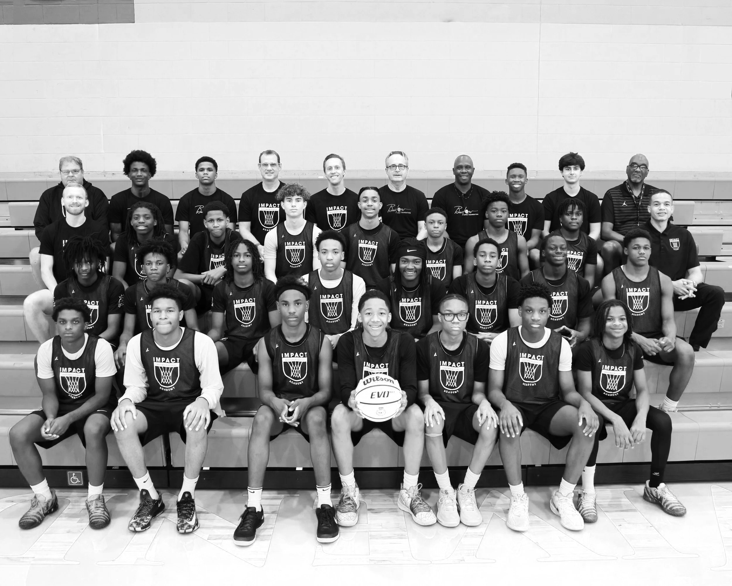 Group of young basketball players and coaches posing for a photo in a gymnasium, some players wearing team jerseys with 'IMPACT' and a basketball logo, holding a basketball in front.