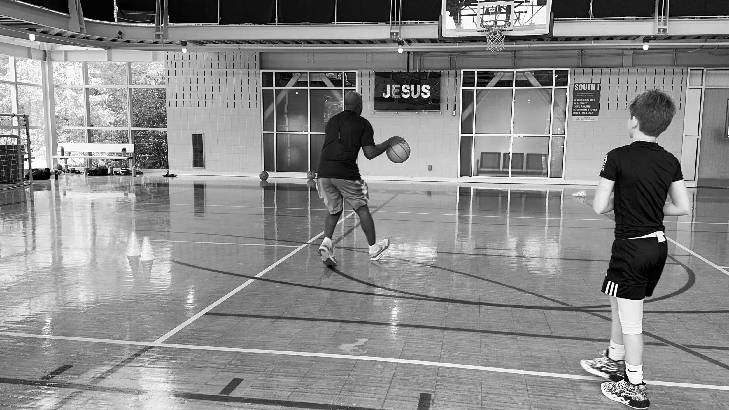 Two boys playing basketball in an indoor court. One boy is dribbling the ball while the other watches. The court has basketball hoops and a media screen displaying the word 'JESUS'.