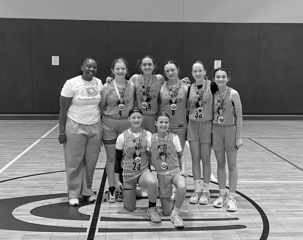 A girls' basketball team of seven players and their coach, all wearing medals, posing on a basketball court. The players are in uniform, and the coach is dressed in casual clothing. All are smiling, and the team appears to have won a game or tournament.