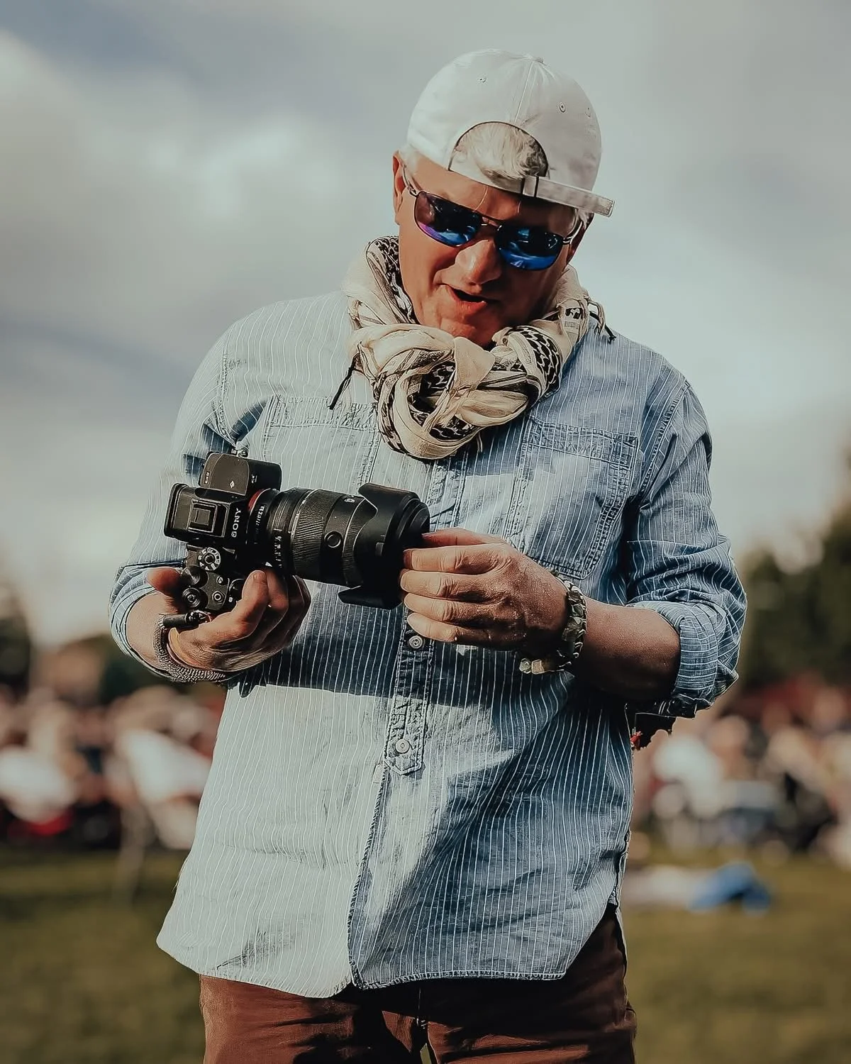 A man wearing a white cap, sunglasses, a striped shirt, and a scarf, holding a professional camera and looking at it with a smile at an outdoor event.