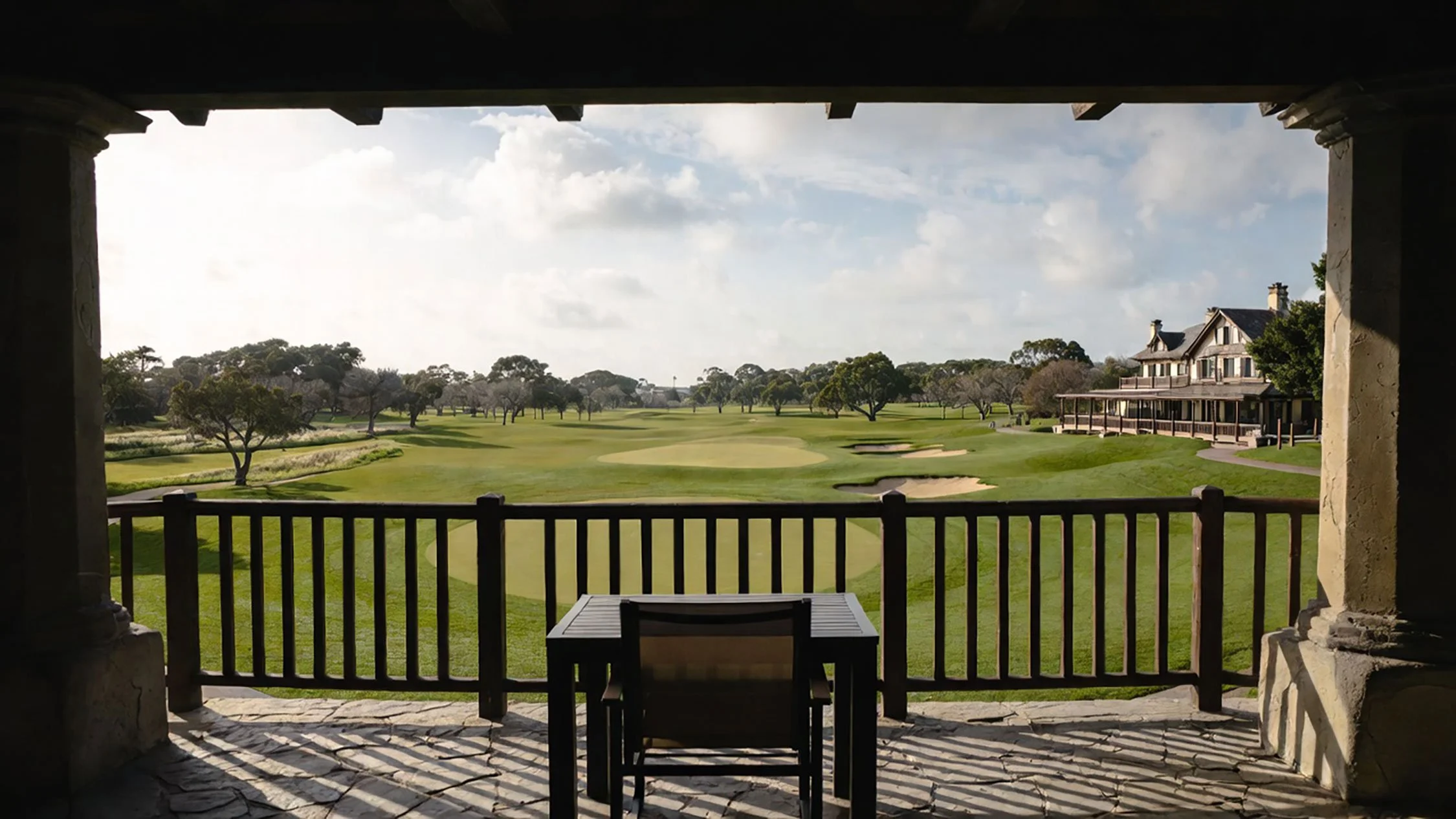 A chair and table on a shaded stone terrace overlooking a golf course and clubhouse.